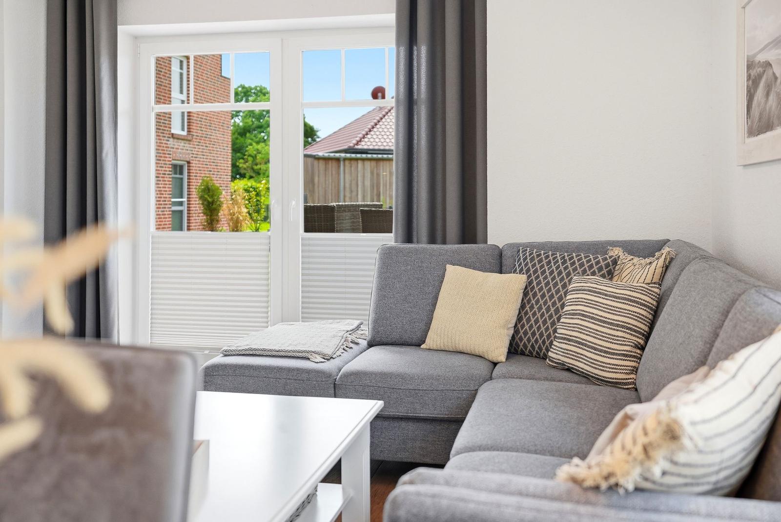 Grauer Sofa-Set mit Kissen und Tisch. Durch Fenster Blick auf Garten und Haus.