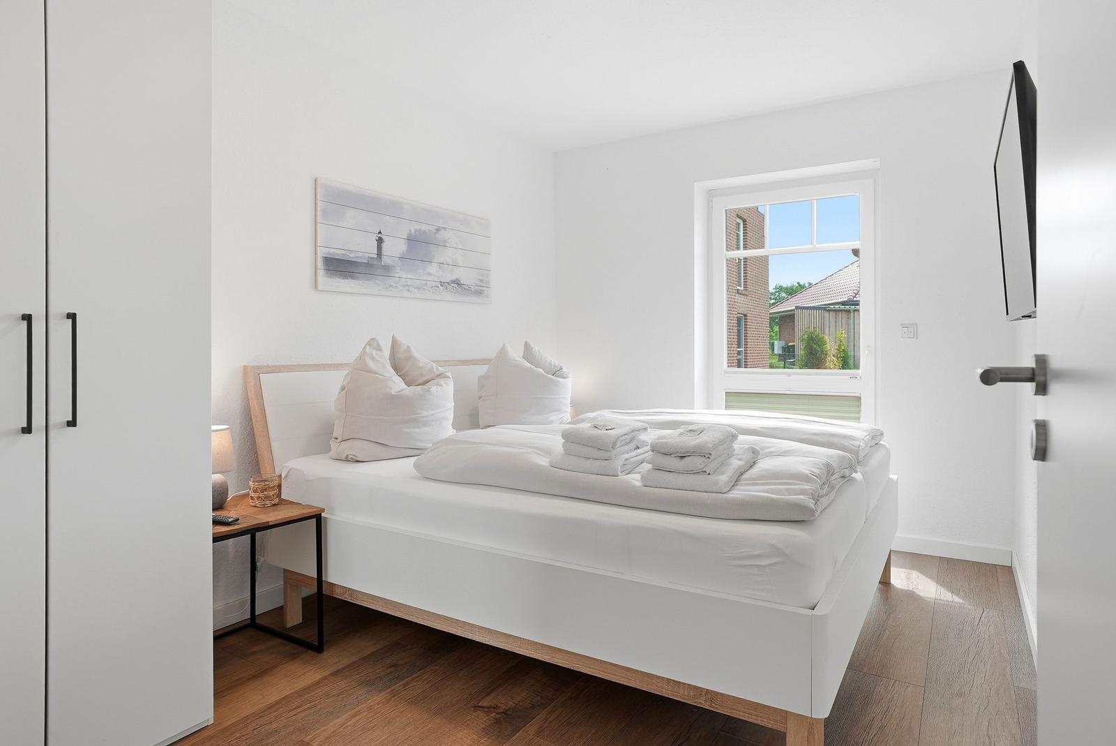 Schlafzimmer mit weißem Bett, Holzboden und Fensterblick auf Außenbereich.