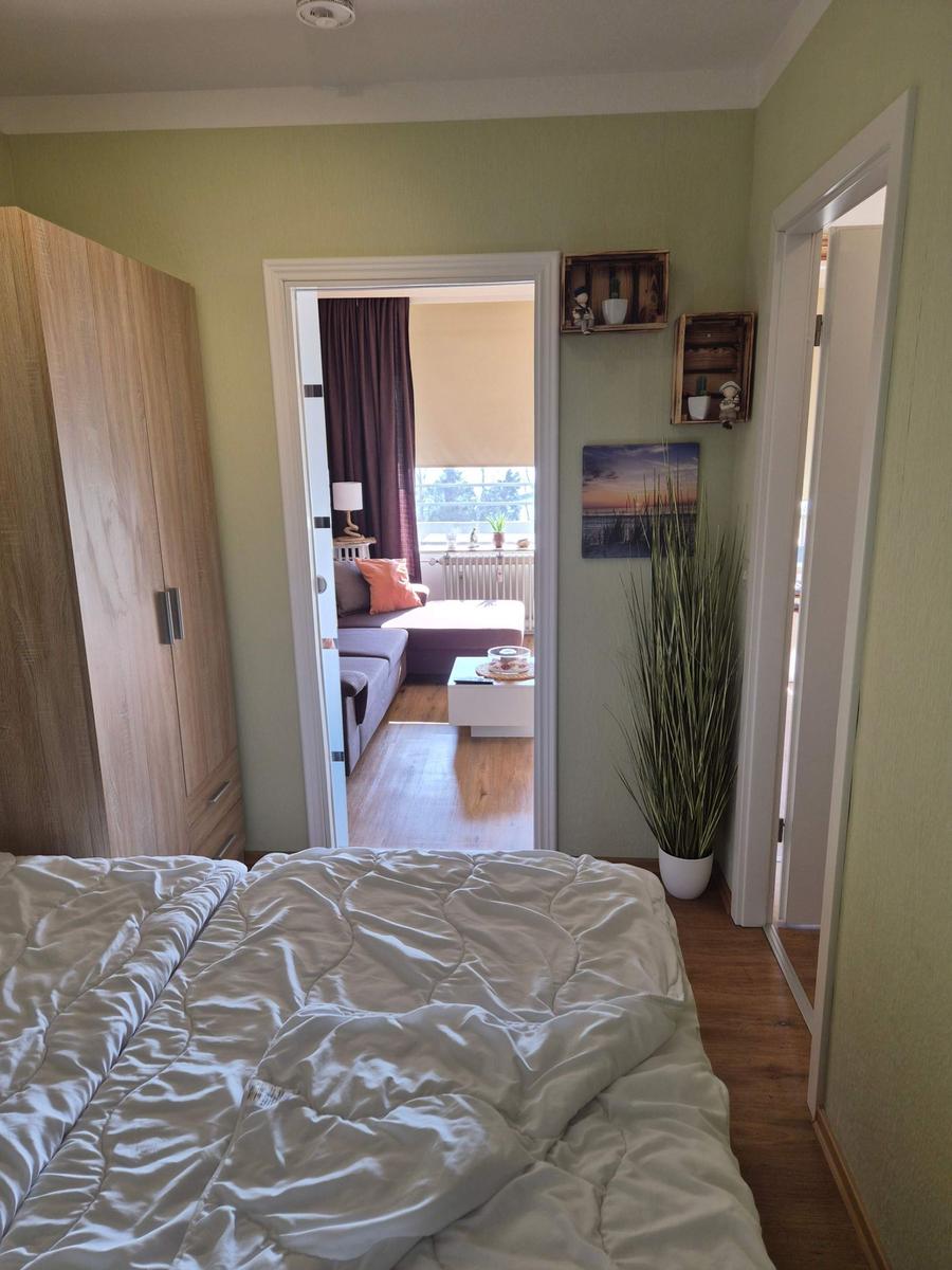 Schlafzimmer mit Blick in Wohnzimmer und Balkon. Holzmöbel und Pflanze sichtbar.