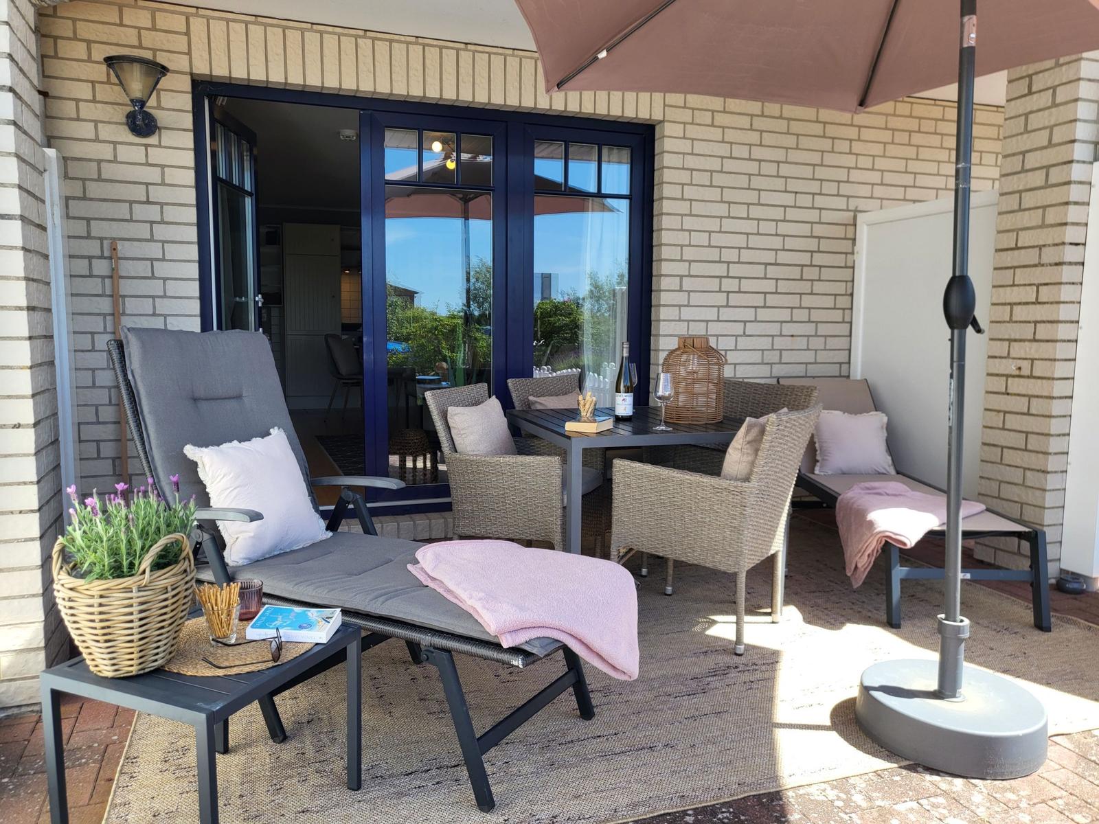 Terrasse mit Liegestuhl, Tisch, Stühlen und Sonnenschirm. Blick in Wohnbereich durch Glastüren.
