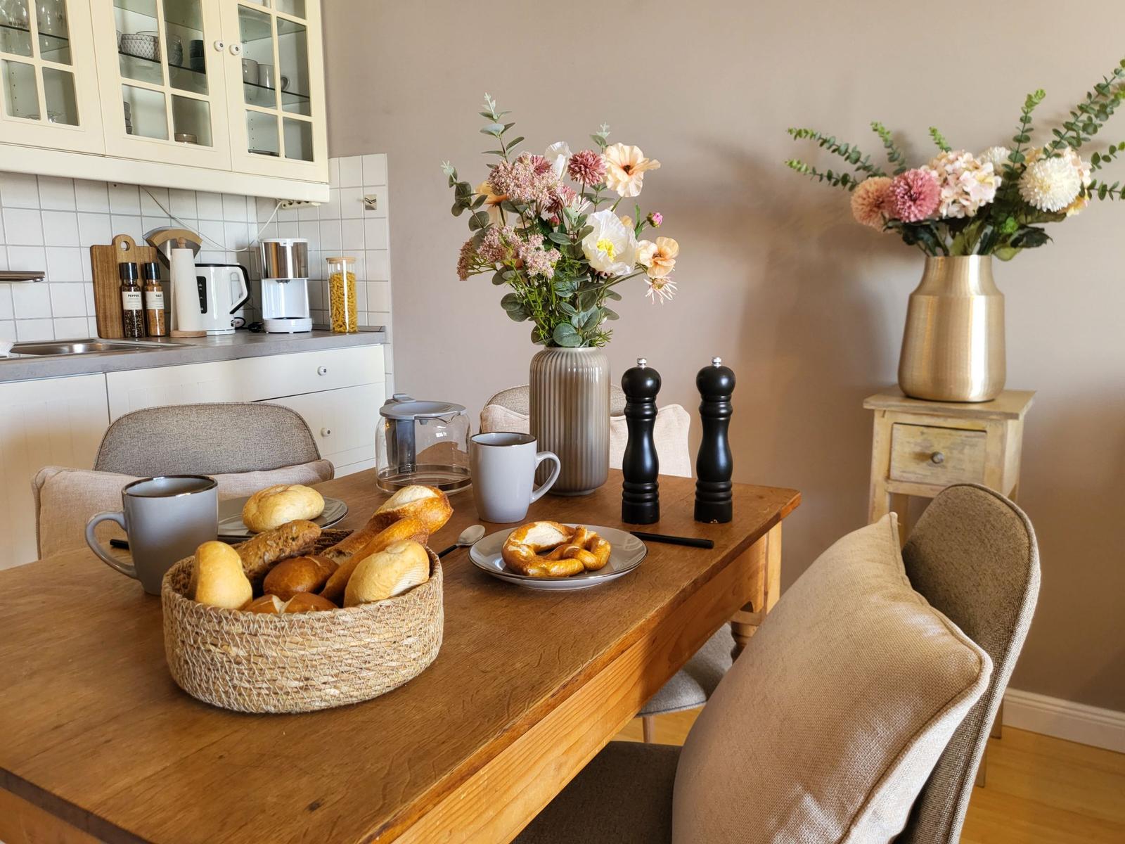 Holztisch mit Brot, Kaffeetassen und Blumenarrangements in Küche und Essbereich.