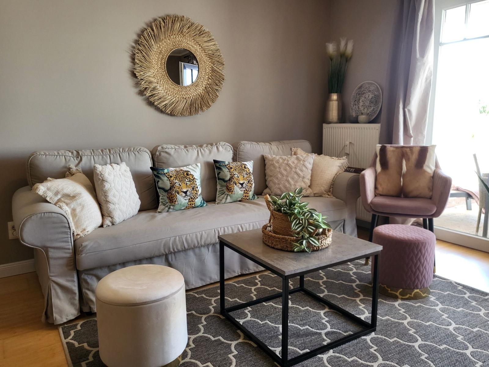 Ein Wohnzimmer mit Sofa, Kissen, Tisch und Stühlen. Wandspiegel und Pflanzen sind sichtbar.