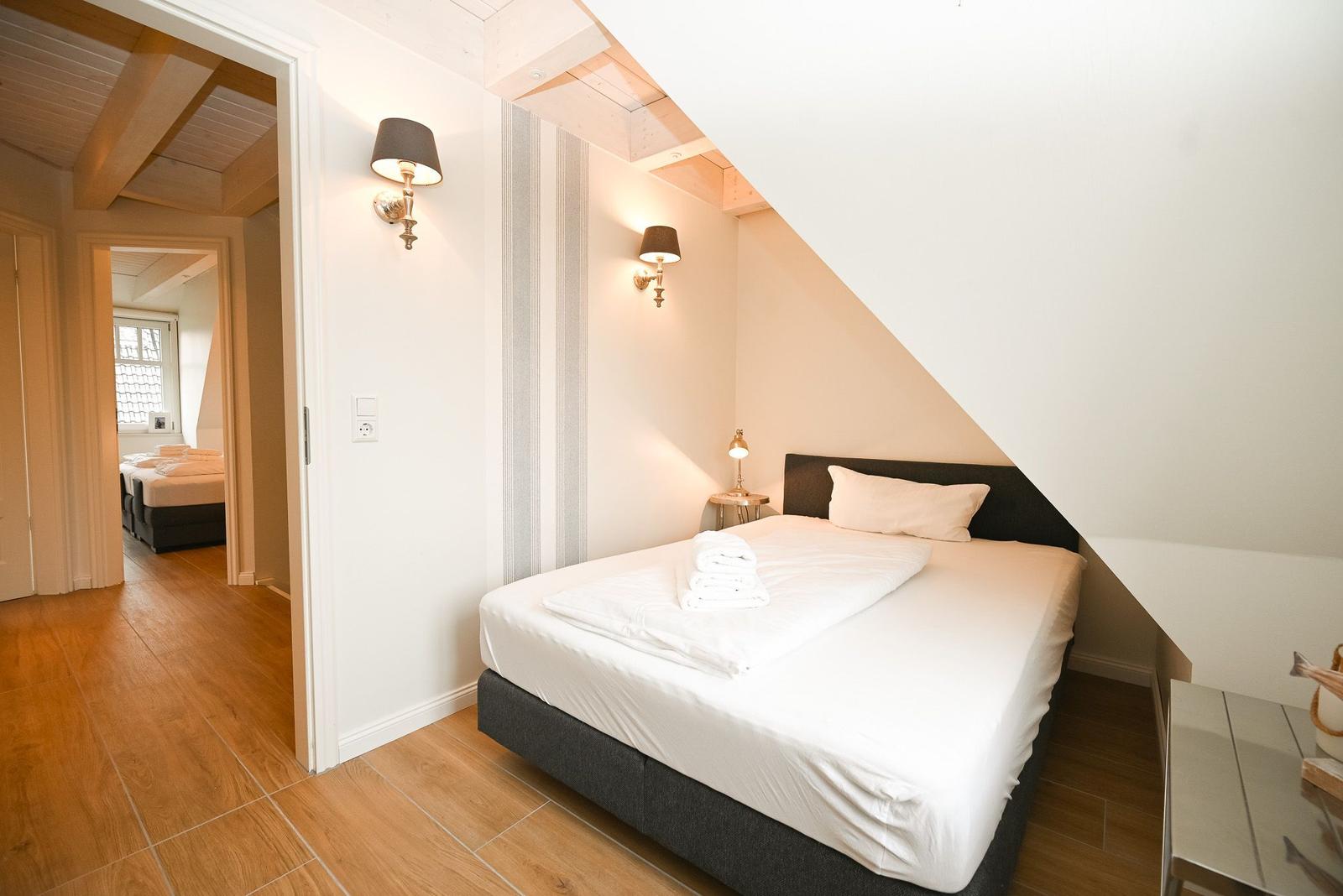 Schlafzimmer mit Dachschräge, Bett, Nachttisch und Stehlampe. Holzboden und Wandbeleuchtung.
