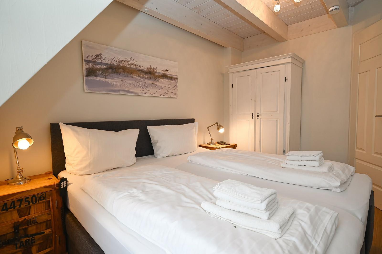 Zimmer mit Doppelbett, weißen Bettwäsche, Nachttisch und Kleiderschrank unter Holzdecke.