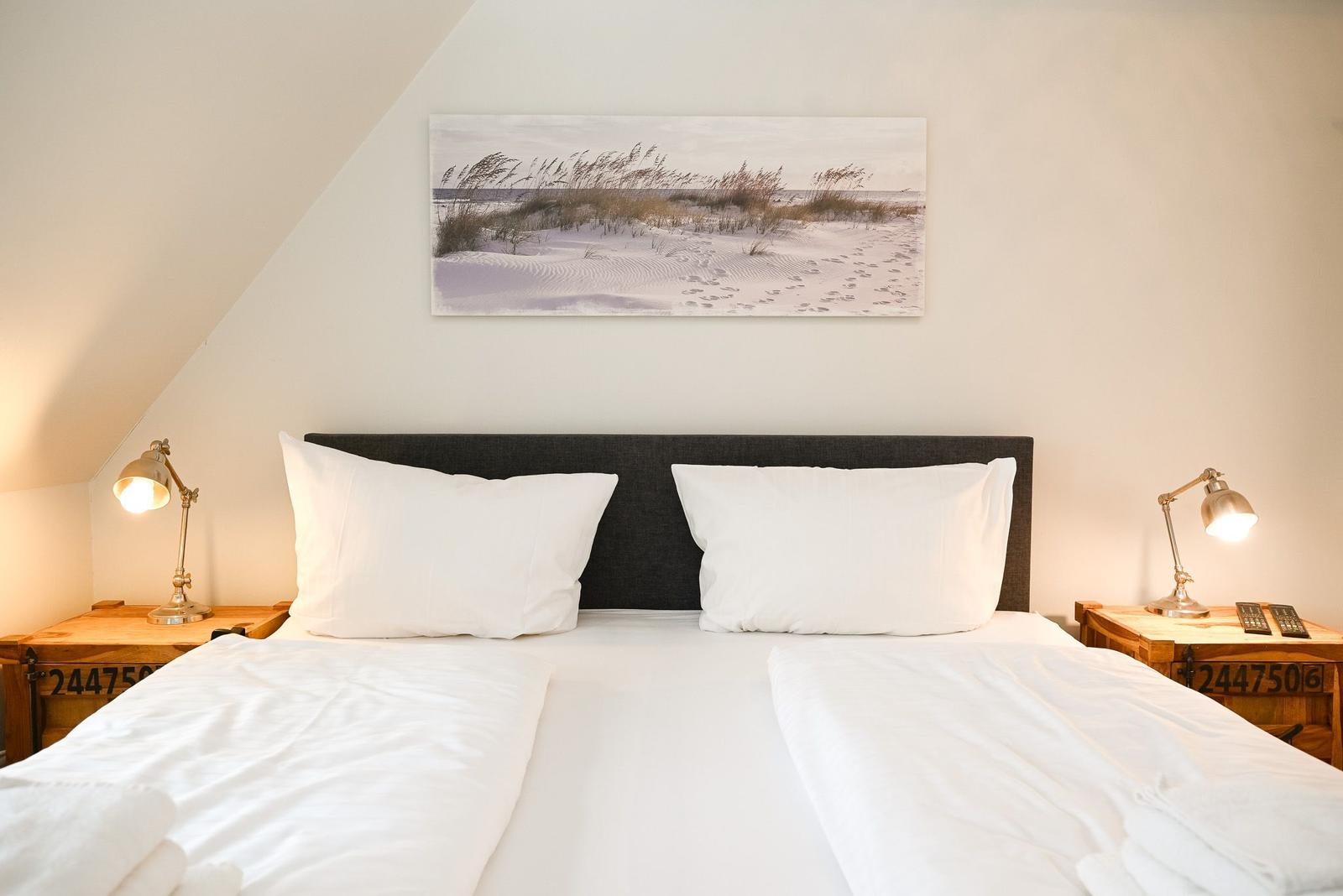Doppelbett mit weißen Laken, zwei Nachttische und Wandbild mit Strandmotiv.