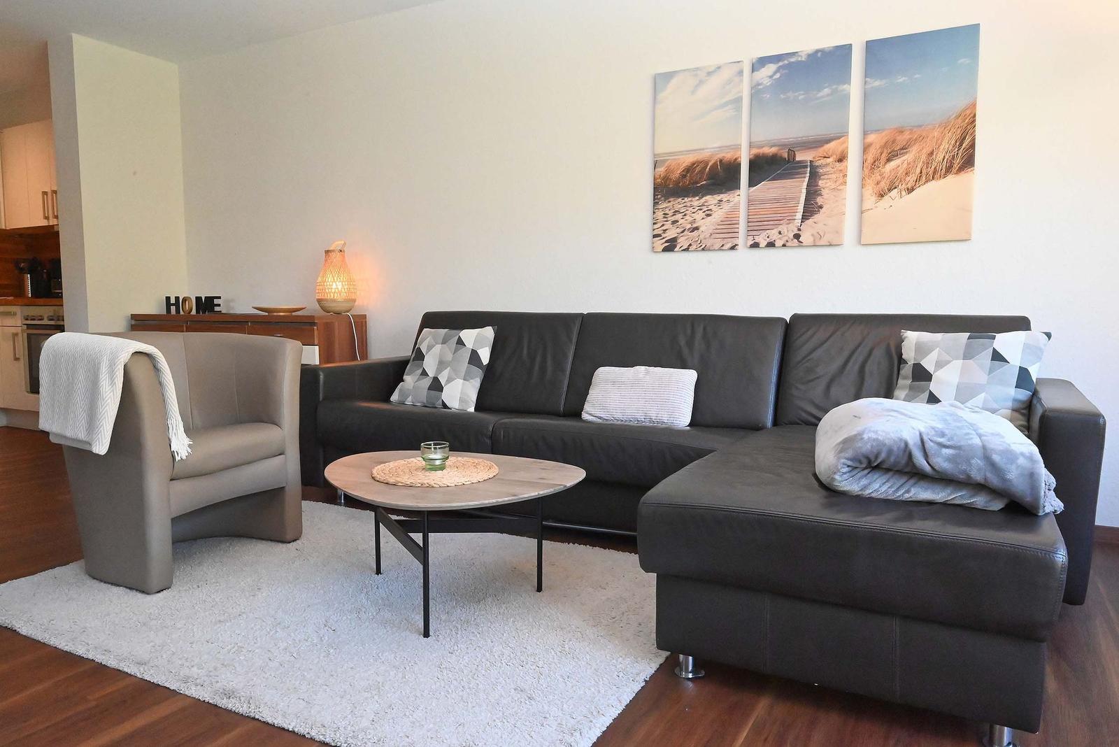 Wohnzimmer mit Sofa, Sessel und Wohnwand mit Strandbildern.