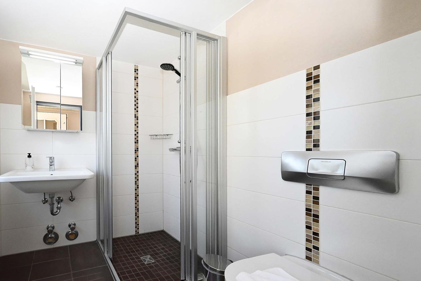 Badezimmer mit Dusche, Waschbecken und Toilette. Fliesen und Spiegel sind sichtbar.