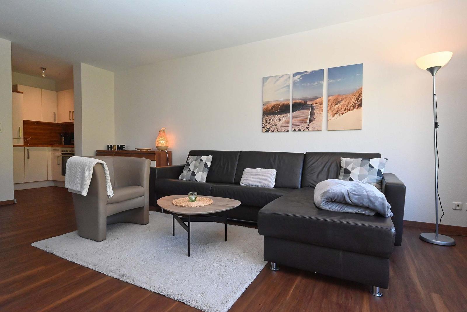 Wohnzimmer mit Sofa, Sessel und Küchenbereich im Hintergrund.