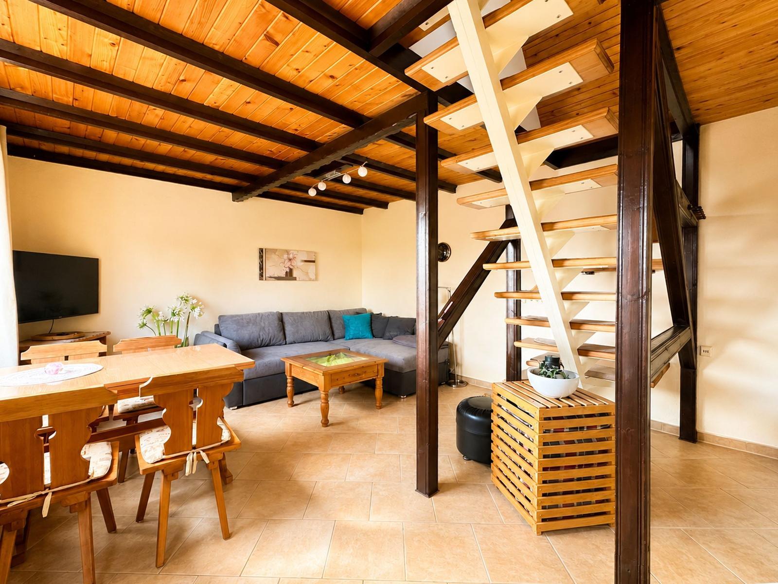 Wohn- und Essbereich mit Holzdecke, Sofa, Tisch und Treppe zur oberen Etage.