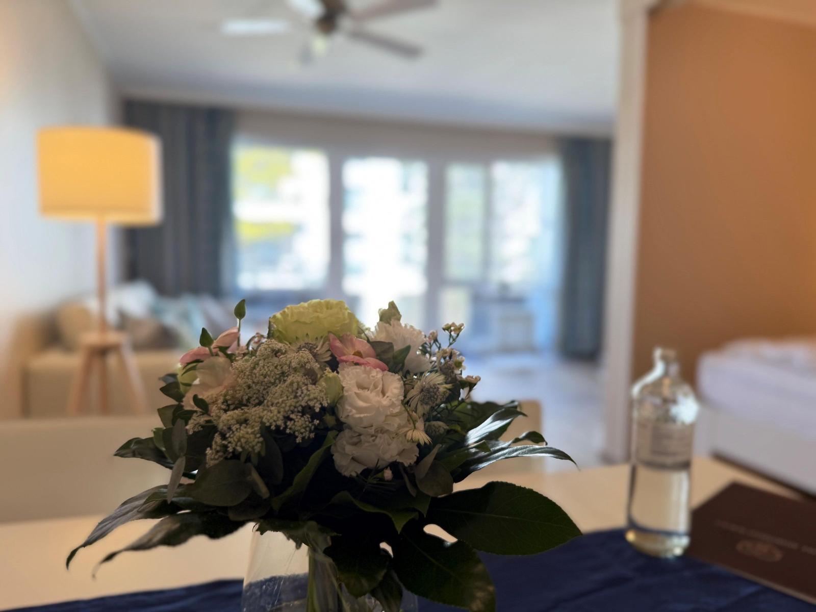 Blumenstrauß auf Tisch, im Hintergrund Wohn- und Schlafbereich mit Fenster.