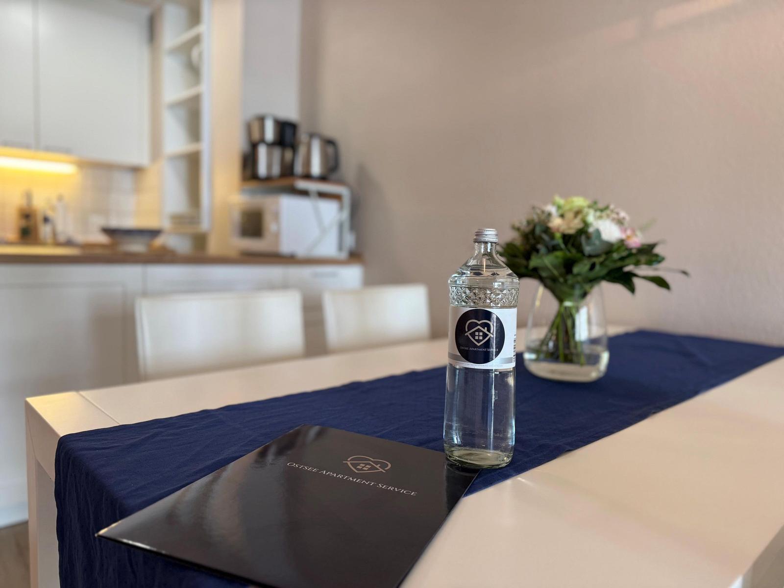 Tisch mit Blumen, Wasserflasche und Broschüre im Essbereich.