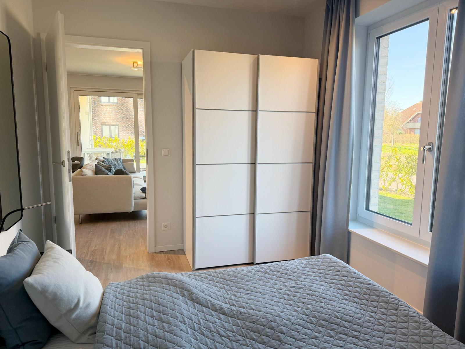 Schlafzimmer mit Bett, Kleiderschrank und Fenster. Durchgang zum Wohnbereich mit Sofa.