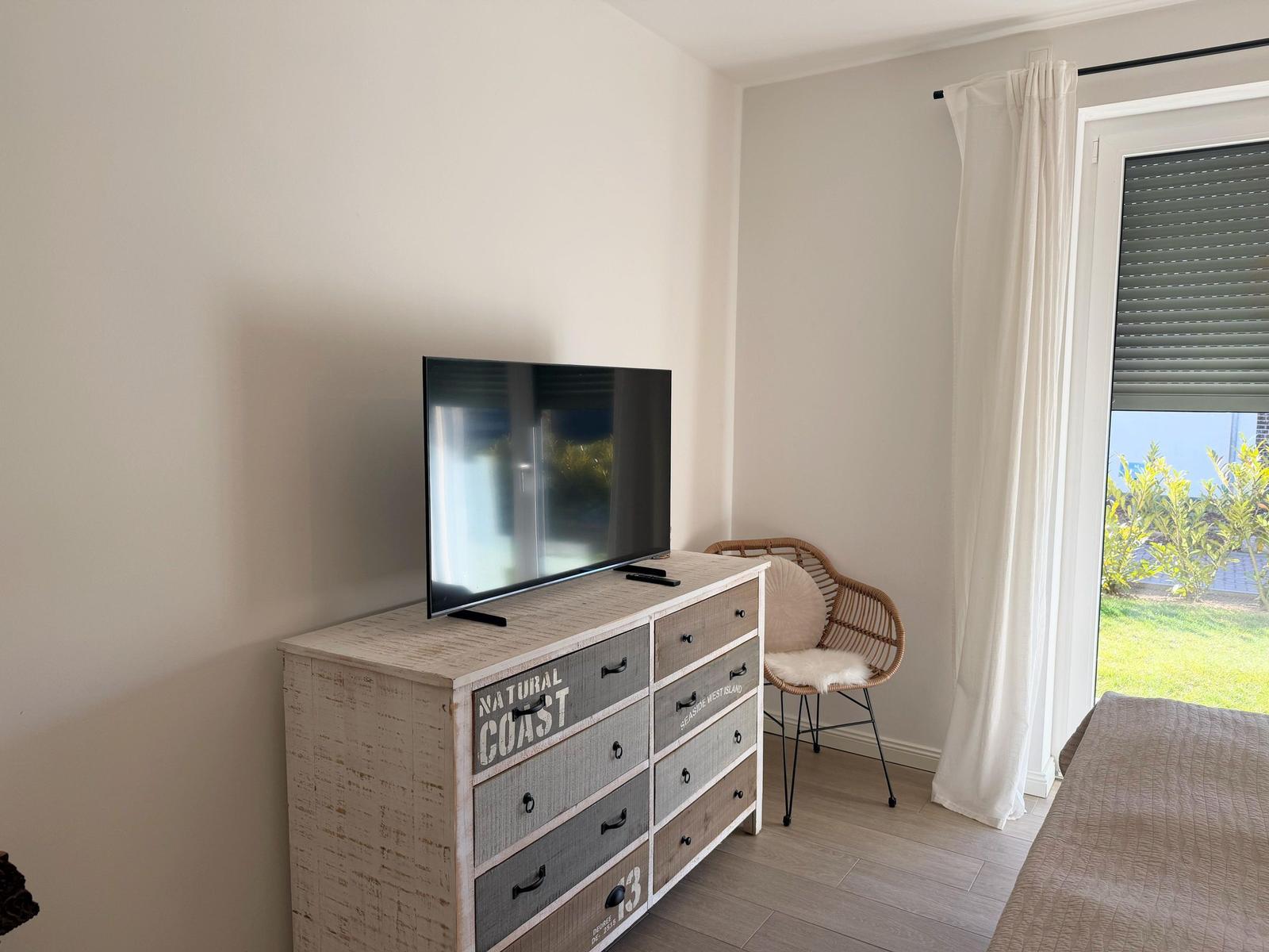 Schlafzimmer mit TV auf Schrank, Stuhl und Fenster mit Blick auf Garten.