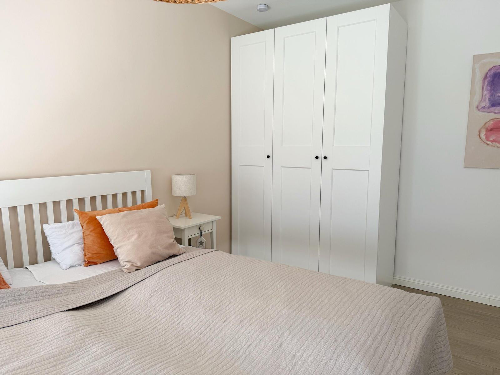 Schlafzimmer mit weißem Bett, Kleiderschrank und Nachttisch mit Lampe.