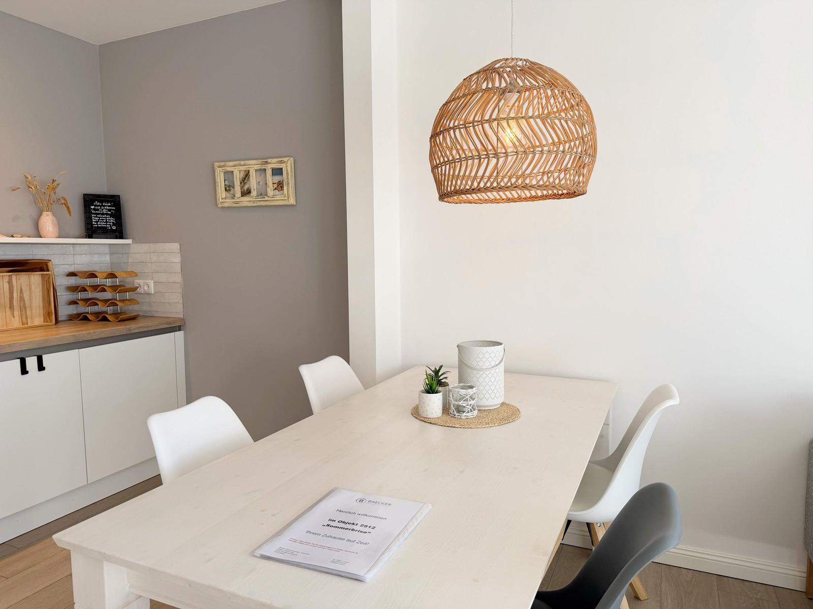 Esstisch mit vier Stühlen unter einer Rattanlampe. Küchenbereich im Hintergrund.