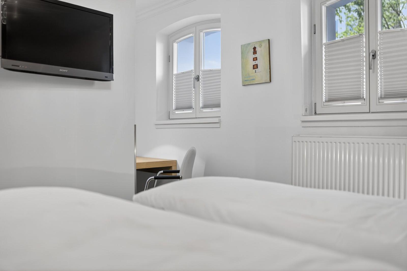 Schlafzimmer mit Bett, Fernseher, Schreibtisch und Fenstern mit Jalousien.