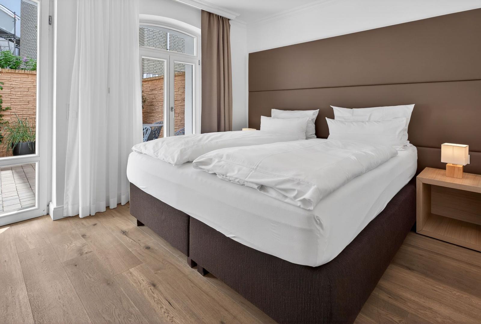 Doppelbett mit weißen Laken, brauner Kopfseite und Holzboden. Durchgang zur Terrasse mit weißen Vorhängen.