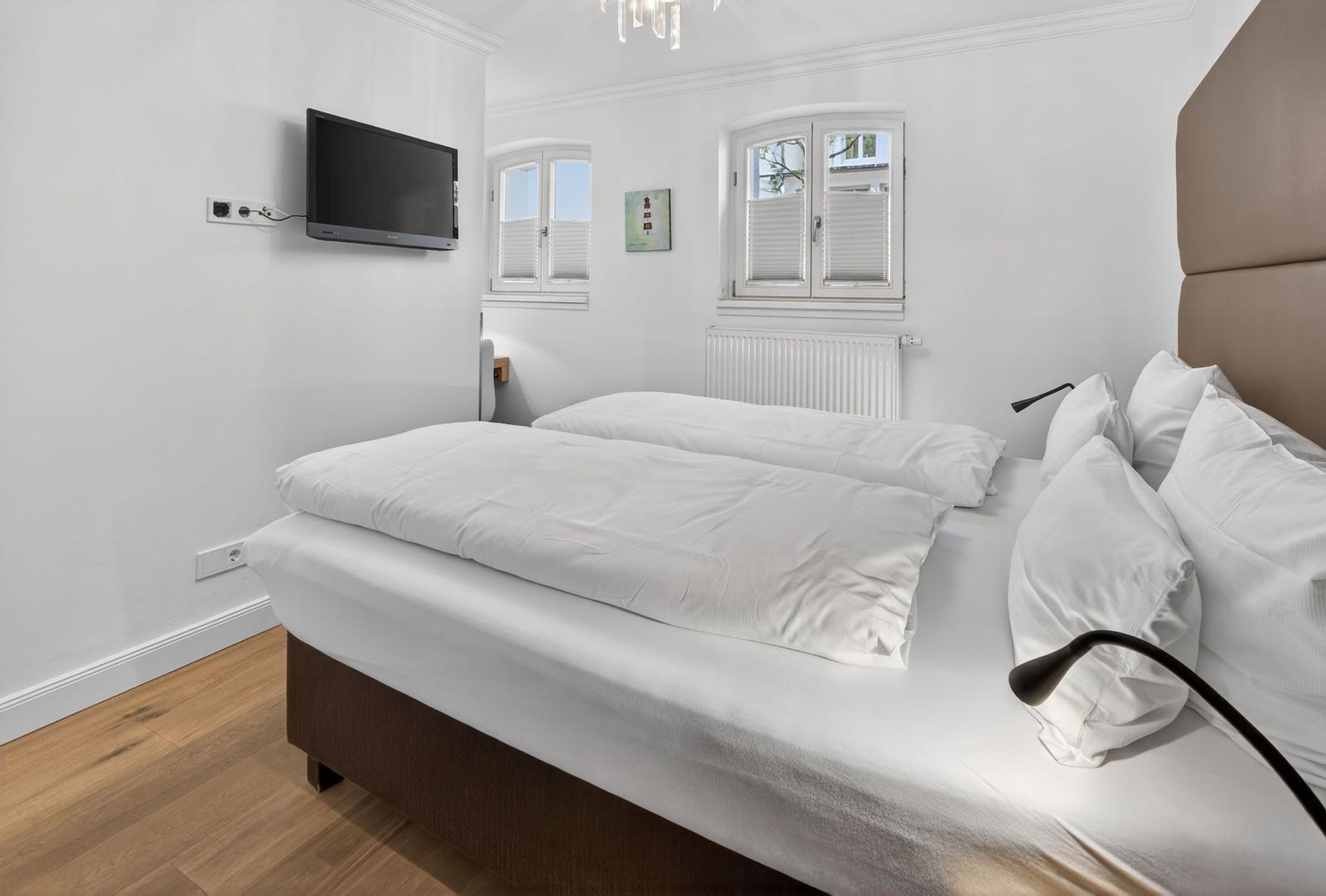 Schlafzimmer mit Doppelbett, Wand-TV und zwei Fenstern mit Jalousien.