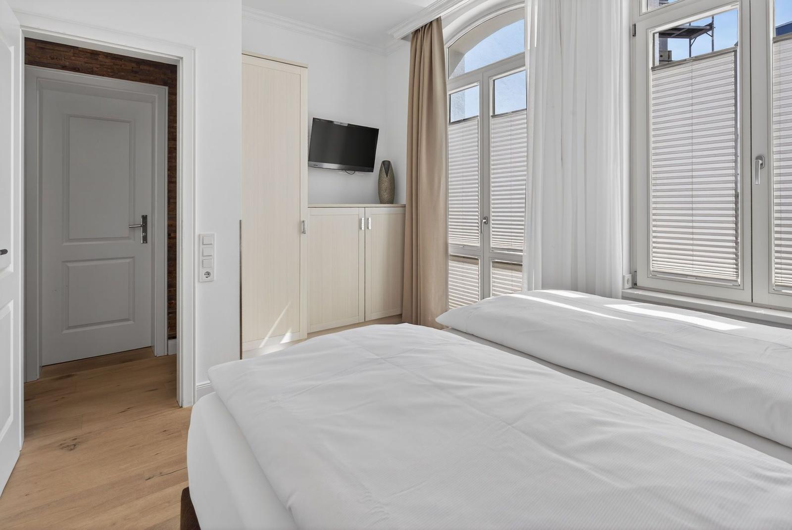 Schlafzimmer mit Bett, Fenster und TV. Holzboden und helle Wände.