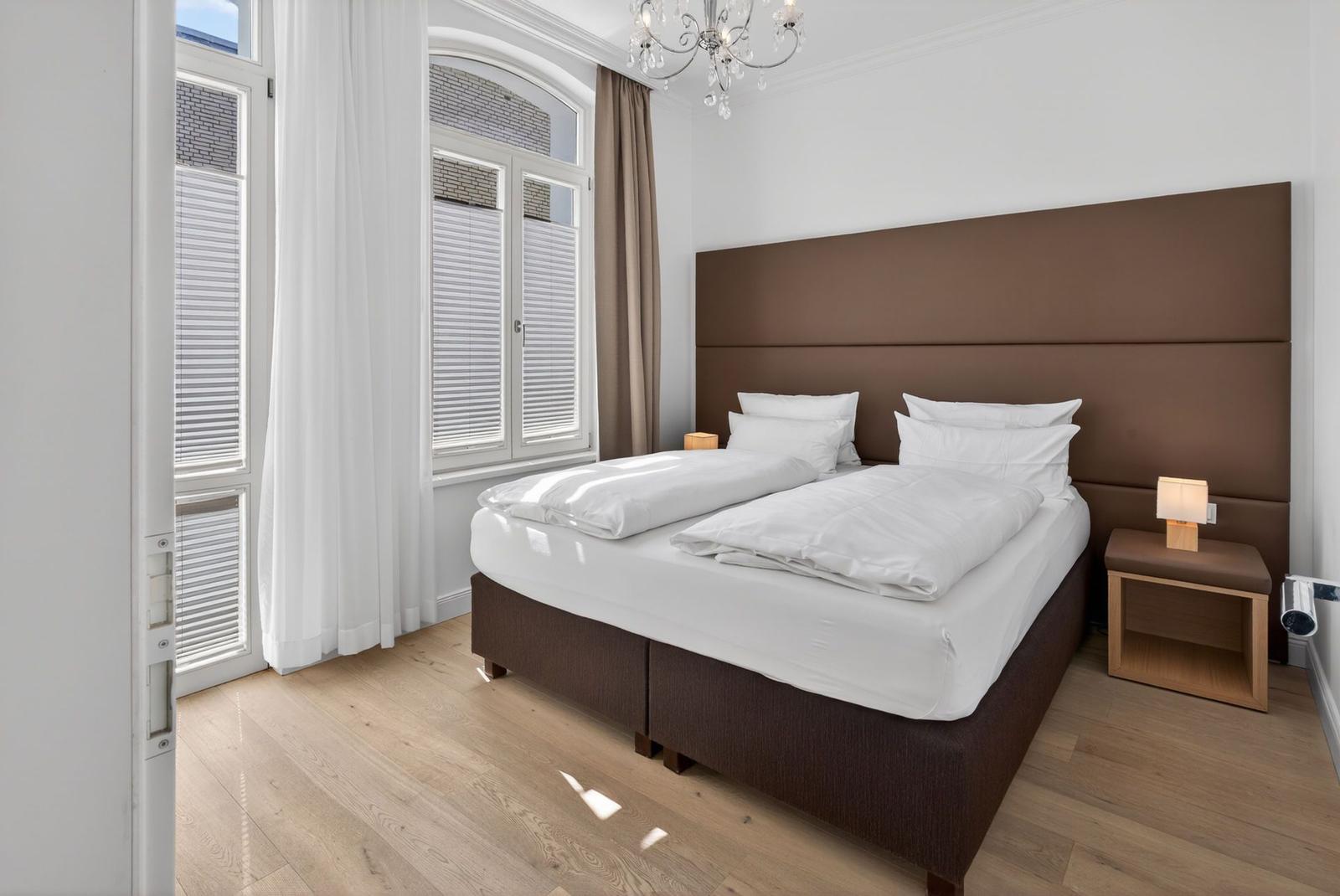 Schlafzimmer mit Doppelbett, braunem Kopfteil, Holzboden und Fenster mit Jalousien.