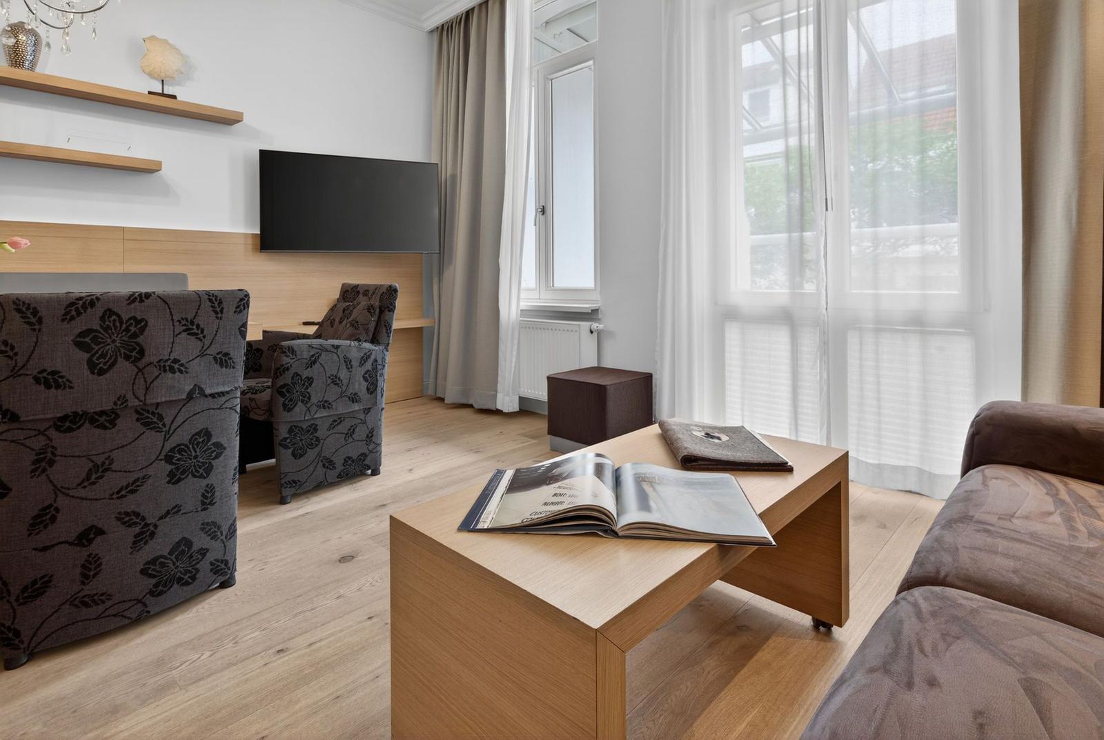 Wohnzimmer mit Sofa, Sessel, Couchtisch und Fernseher. Große Fenster mit Vorhängen.