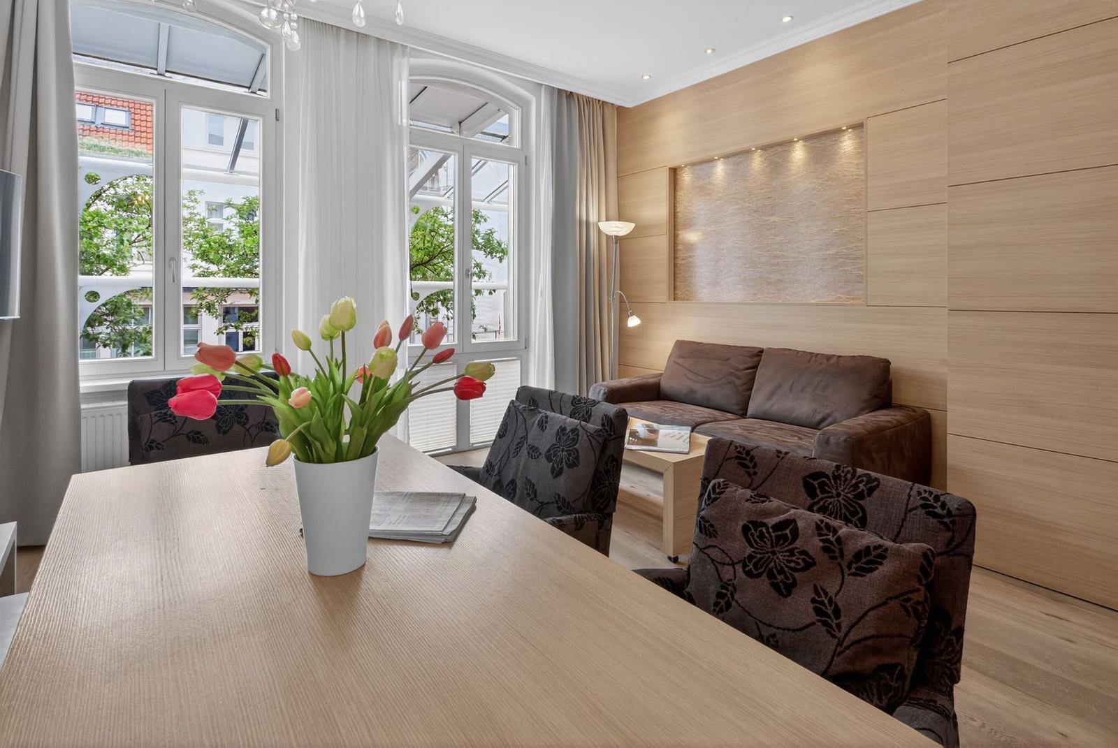 Ein Essbereich mit Tisch und Stühlen. Im Hintergrund ein Sofa und große Fenster.