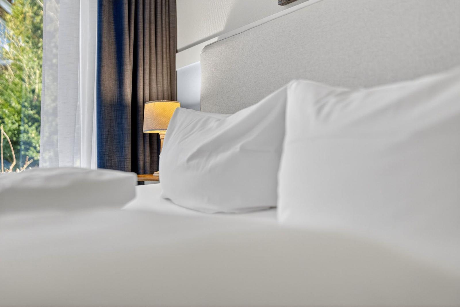Hochwertiges Bett mit weißen Kissen und Kopfteil. Neben einem Nachttisch mit Lampe und Fenster mit Vorhängen.