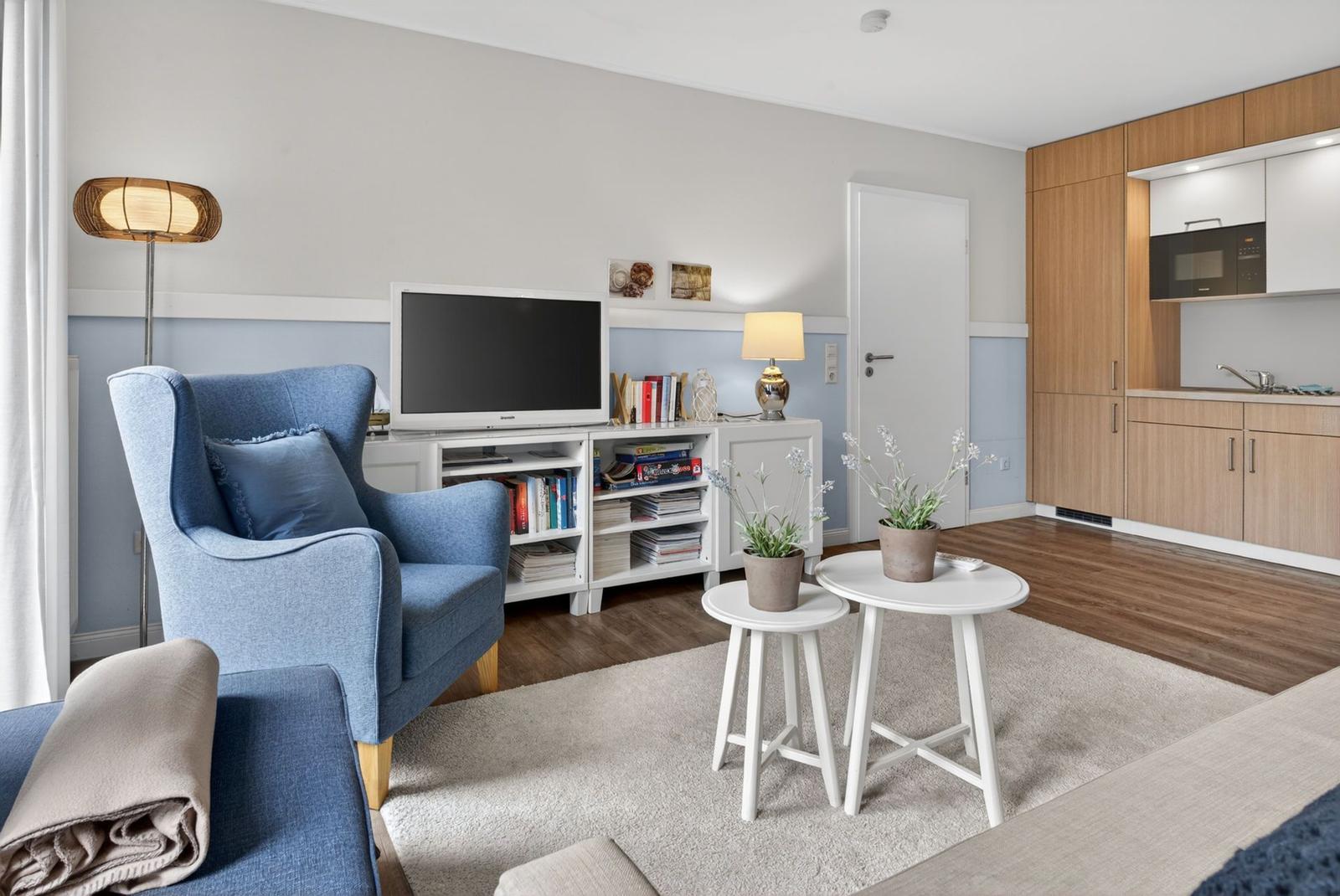Wohnbereich mit blauem Sessel, Fernseher und Küchenzeile mit Holzmöbeln.