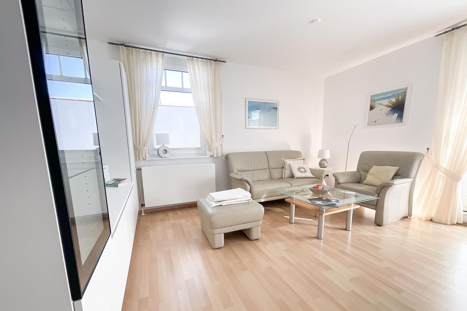 Living room with sofa, armchair, glass table, and wooden floor. Windows with white curtains.