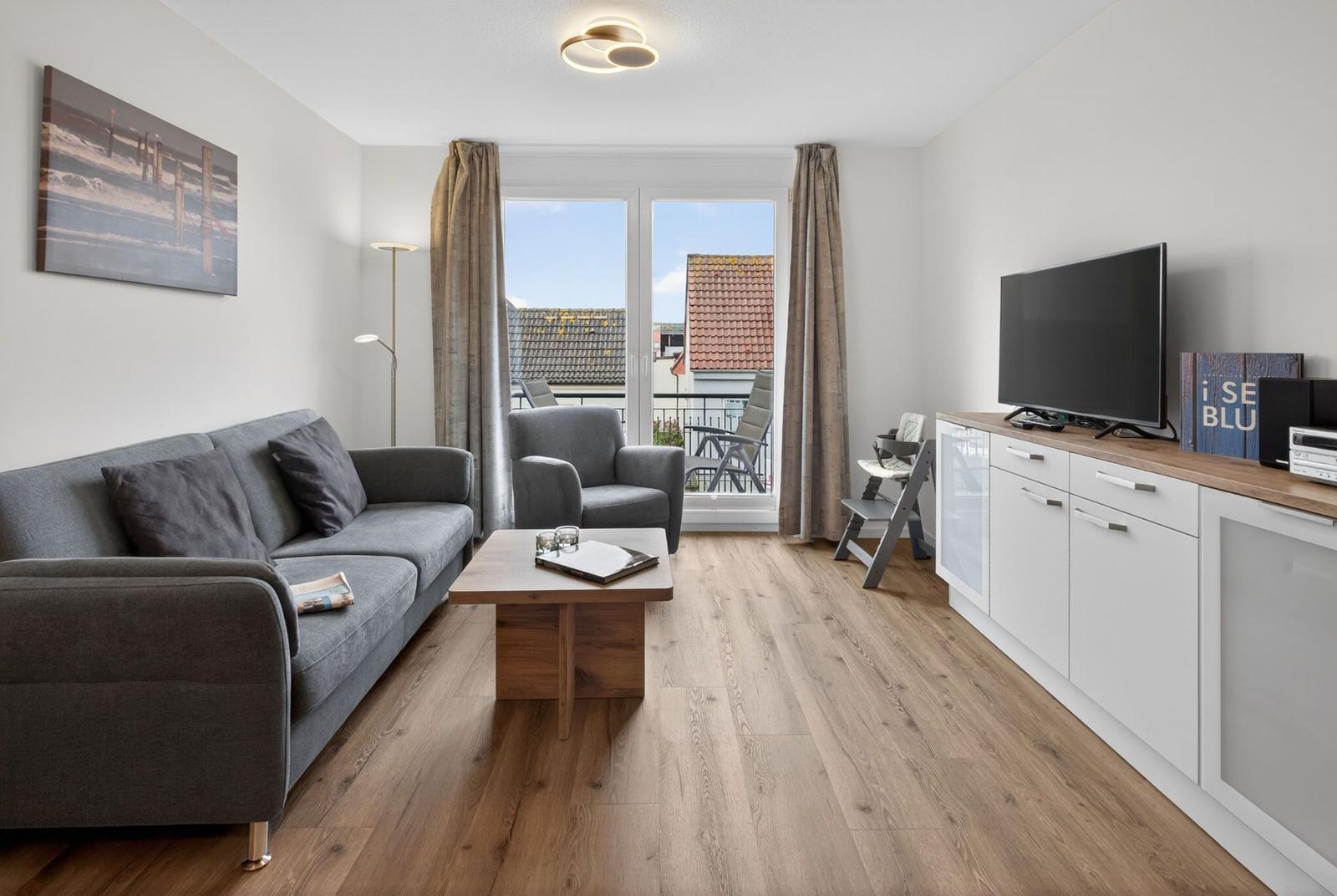 Wohnzimmer mit Sofa, Couchtisch und TV. Balkon mit Sitzgelegenheiten. Holzboden und Wandbild.