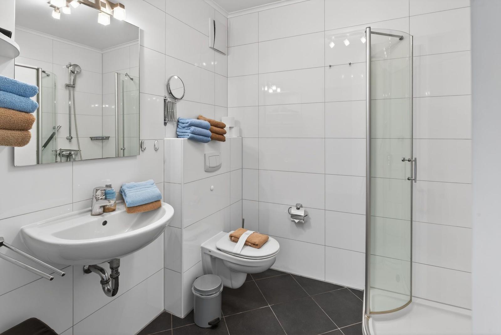 Badezimmer mit Dusche, Waschbecken und Toilette. Weiße Fliesen und Handtücher.