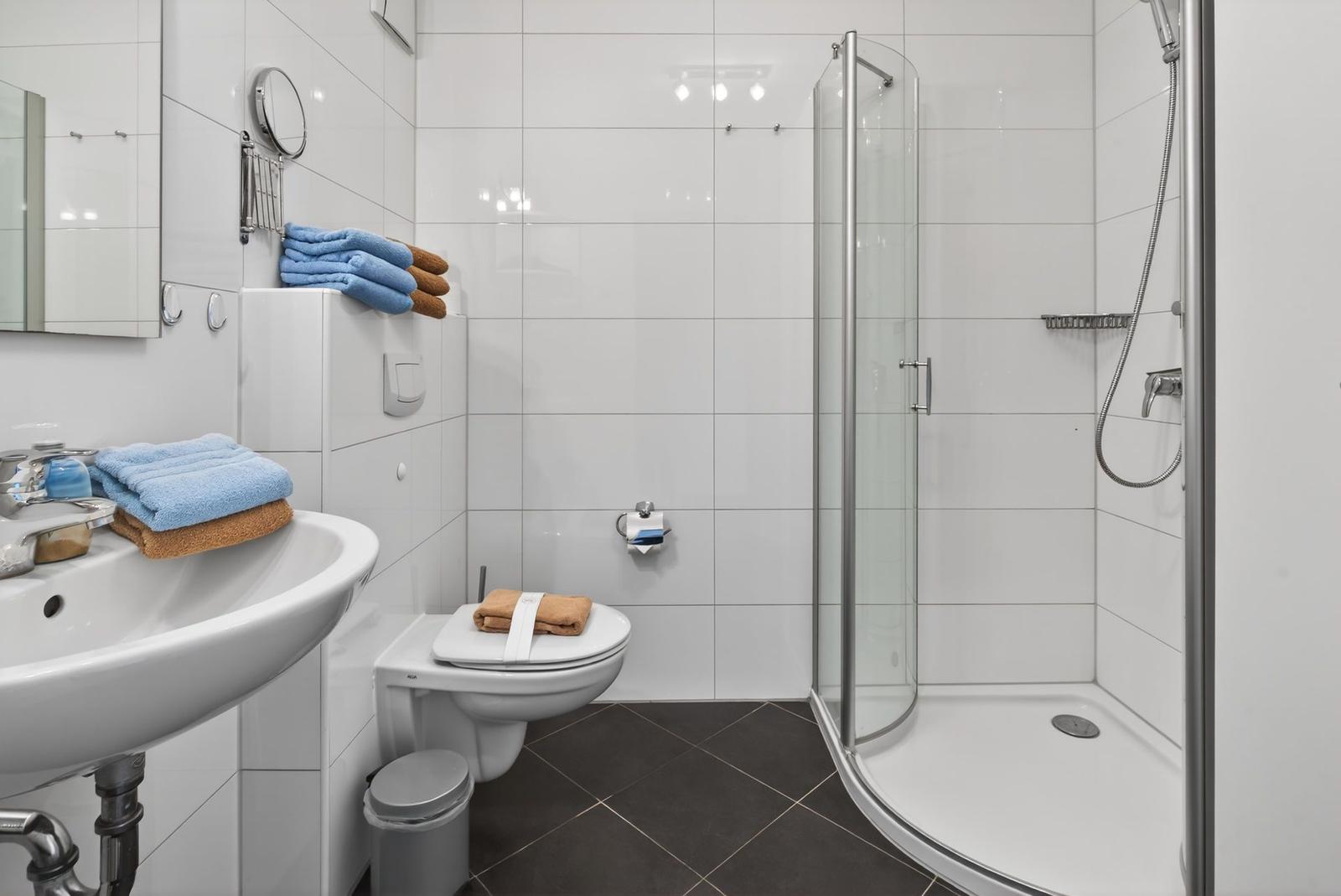 Badezimmer mit Dusche, Toilette und Waschbecken. Weiße Fliesen und braune Bodenfliesen.