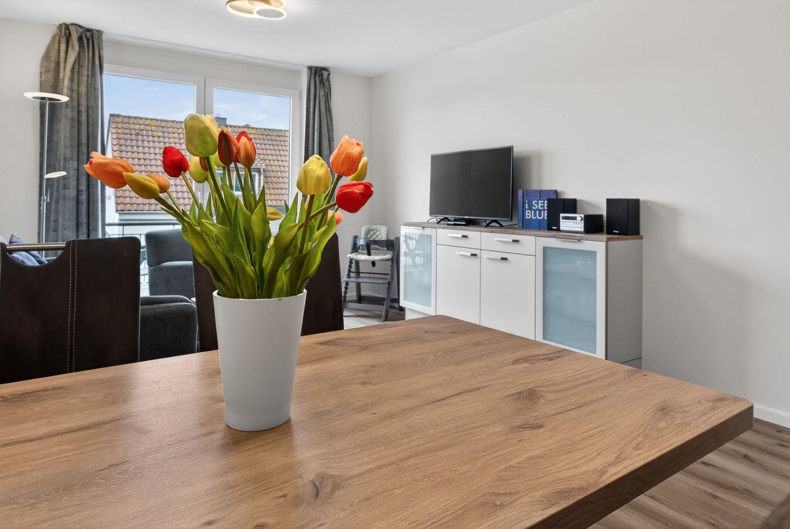 Holztisch mit Tulpenblumen, Fernseher und Küchenzeile im Wohnbereich.