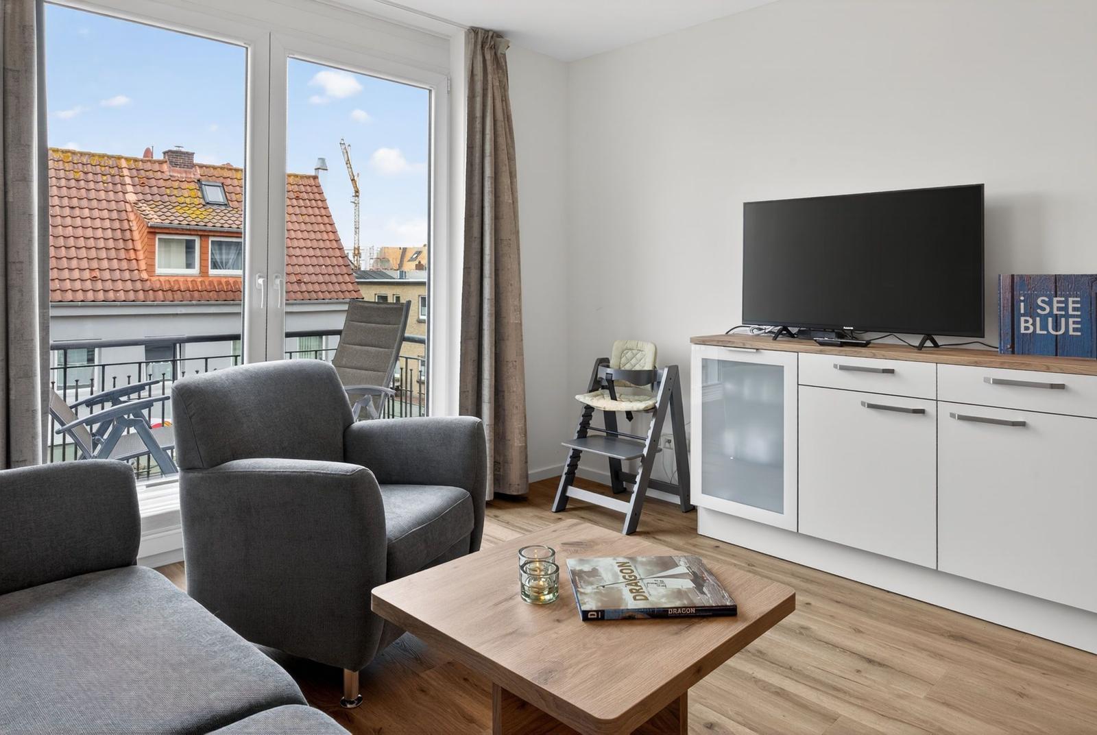 Wohnzimmer mit TV, Sitzgelegenheiten und Balkonblick auf Dächer.