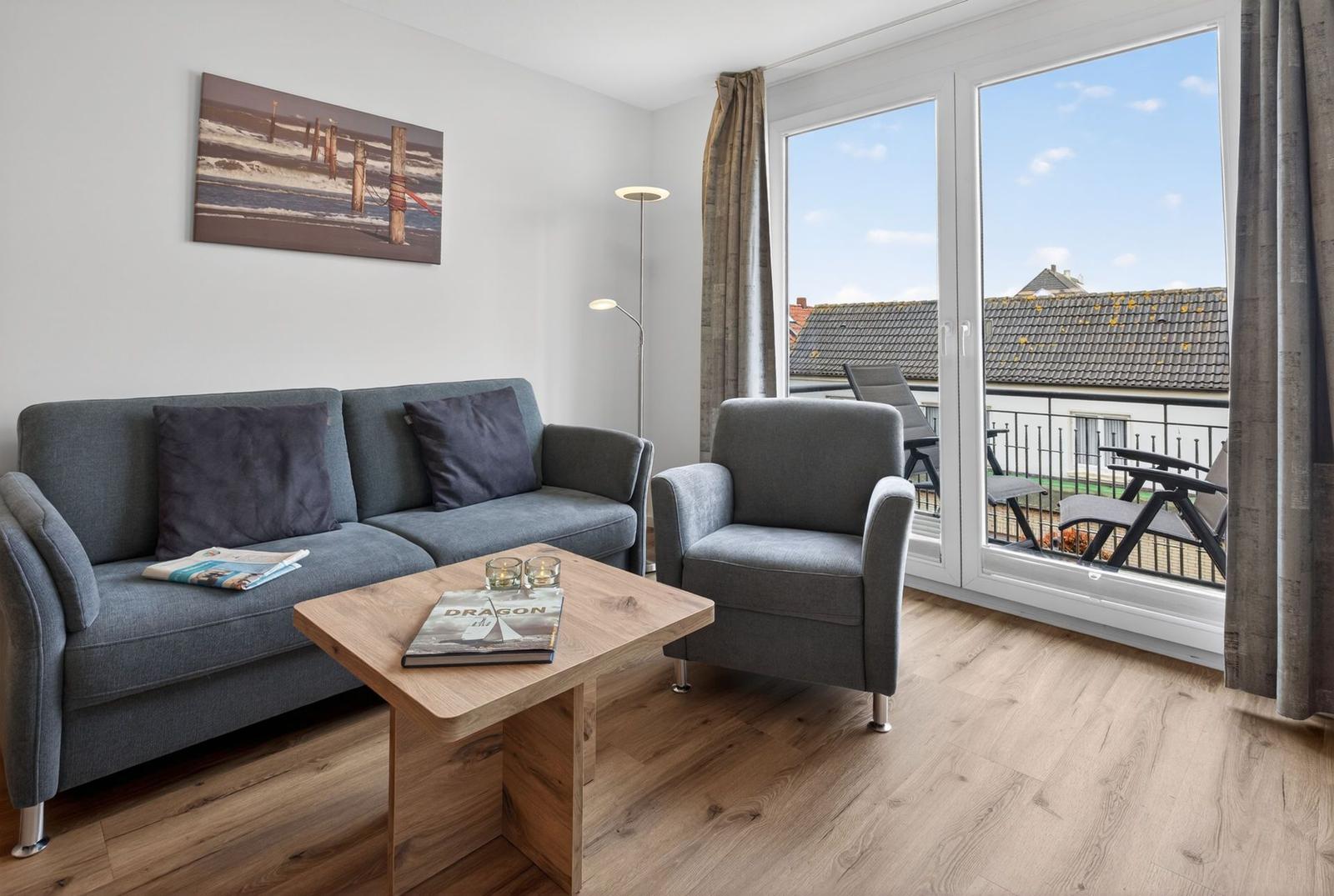Wohnzimmer mit Sofa, Sessel und Balkon. Holztisch und Wandbild sind sichtbar.