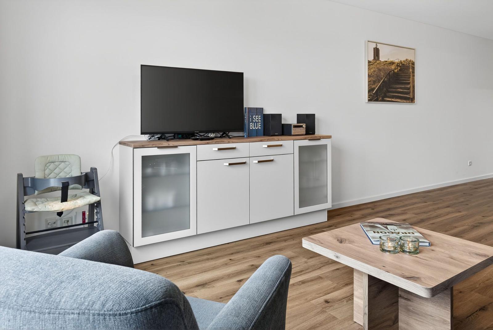 Wohnzimmer mit Fernseher, Sideboard und Holztisch. Hochstuhl und Wandbild sind sichtbar.