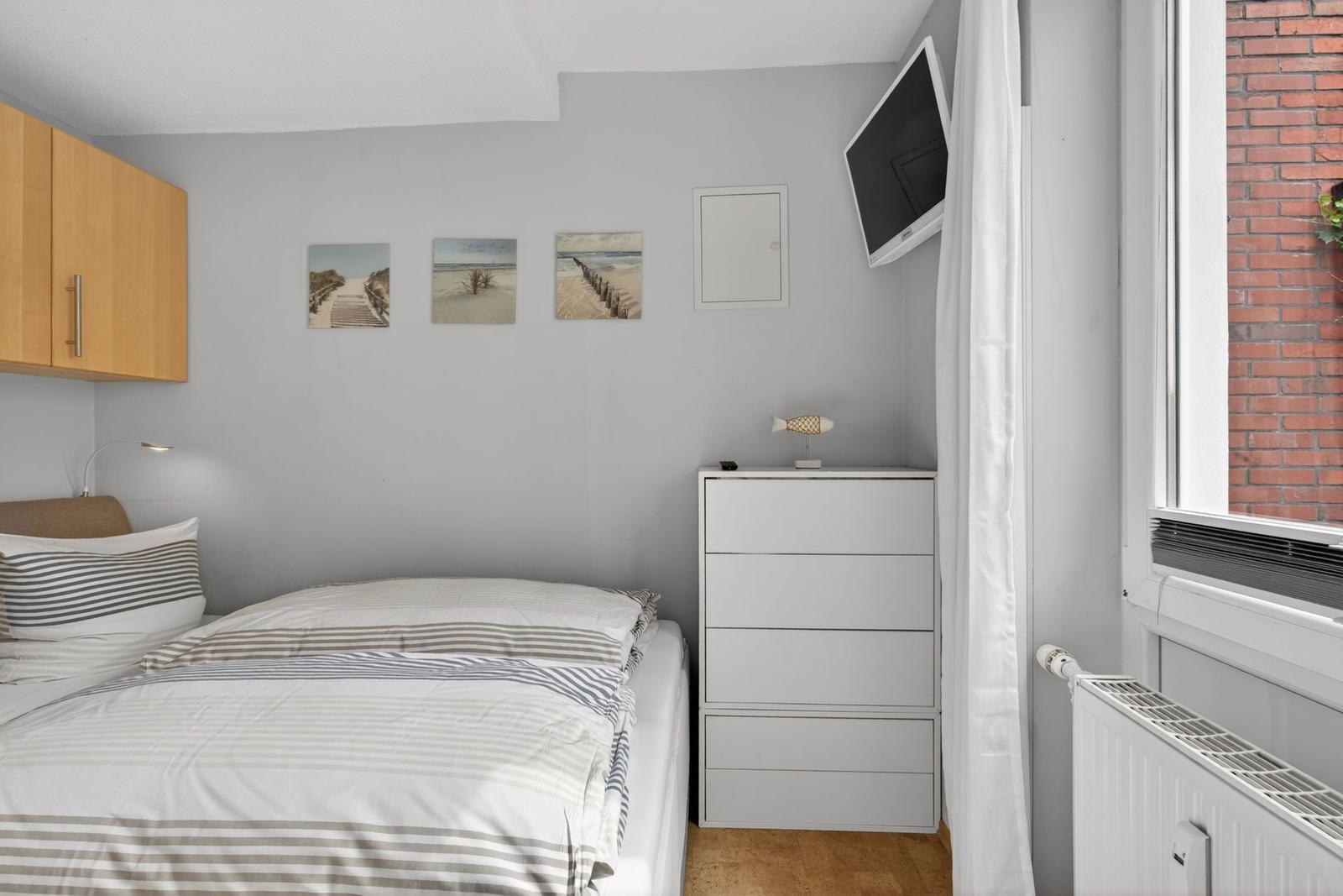Schlafzimmer mit Bett, Schrank und Wandbildern. Fenster mit Blick auf Backsteinmauer.