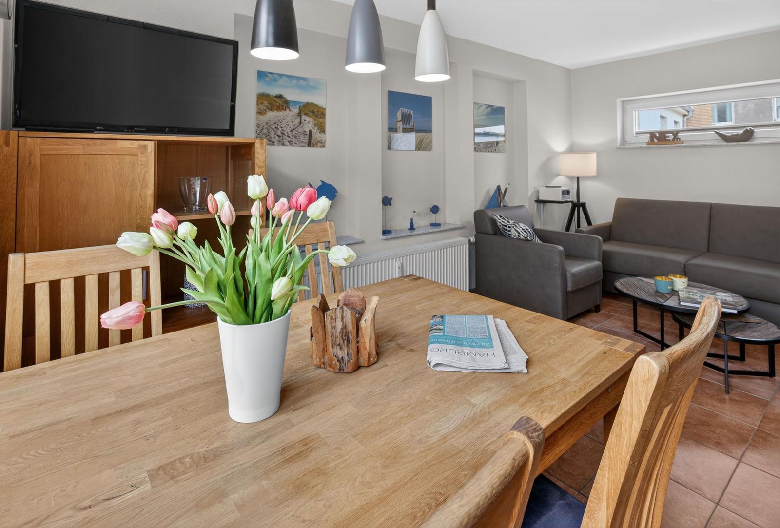 Essbereich mit Holztisch, Blumen und Fernseher. Wohnbereich mit Sofa und Sessel.