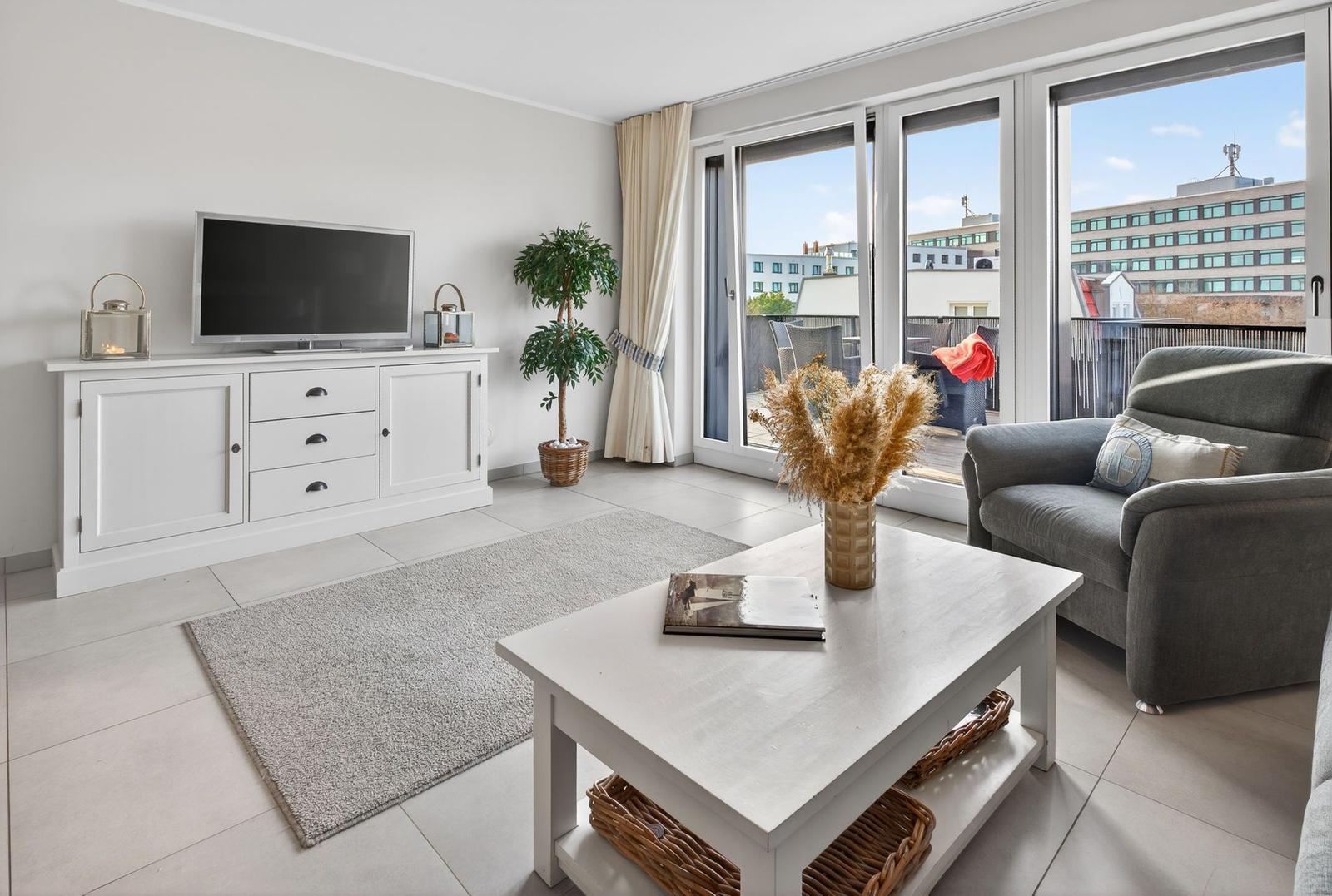 Wohnzimmer mit TV, Couch und Balkon. Fenster öffnen sich zum Stadtblick.