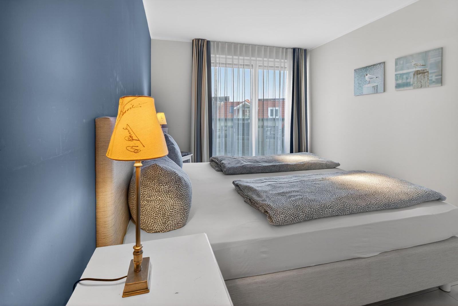 Schlafzimmer mit Bett, Nachttisch und Lampe. Fenster mit Blick auf Gebäude.