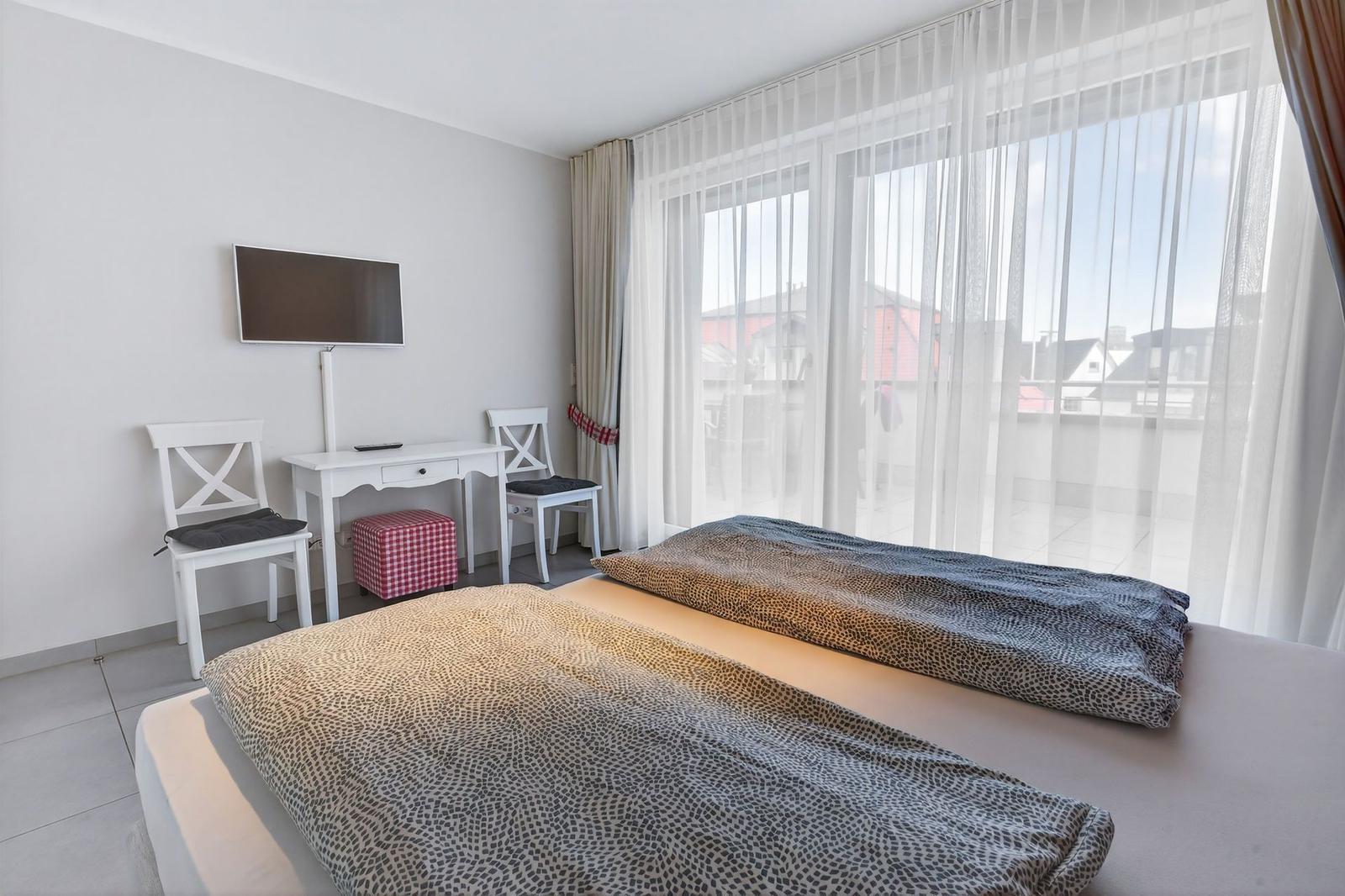 Schlafzimmer mit Bett, Schreibtisch, zwei Stühlen und TV. Große Fenster mit Blick auf Dächer.