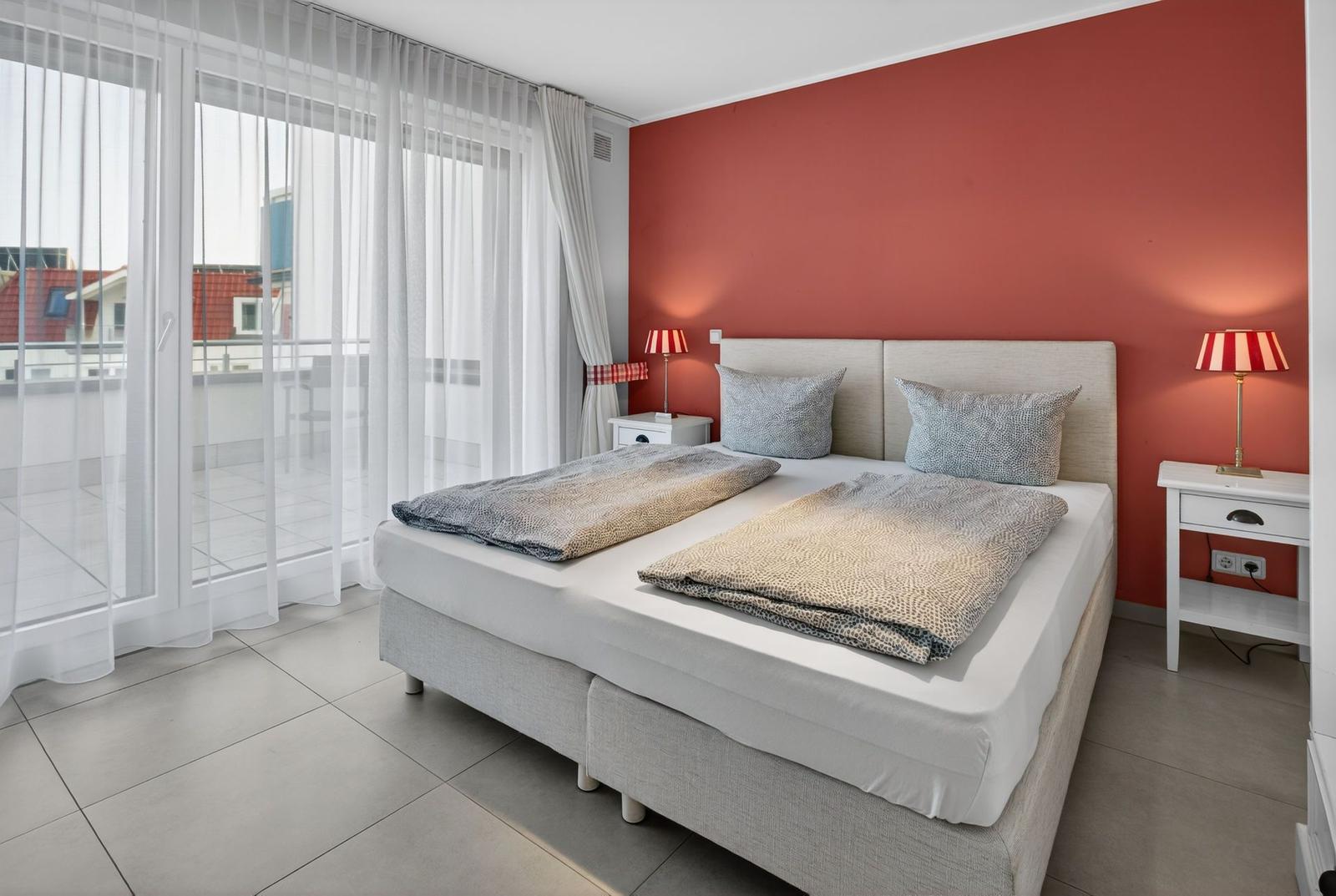 Schlafzimmer mit Doppelbett, roter Wand und Balkontür.