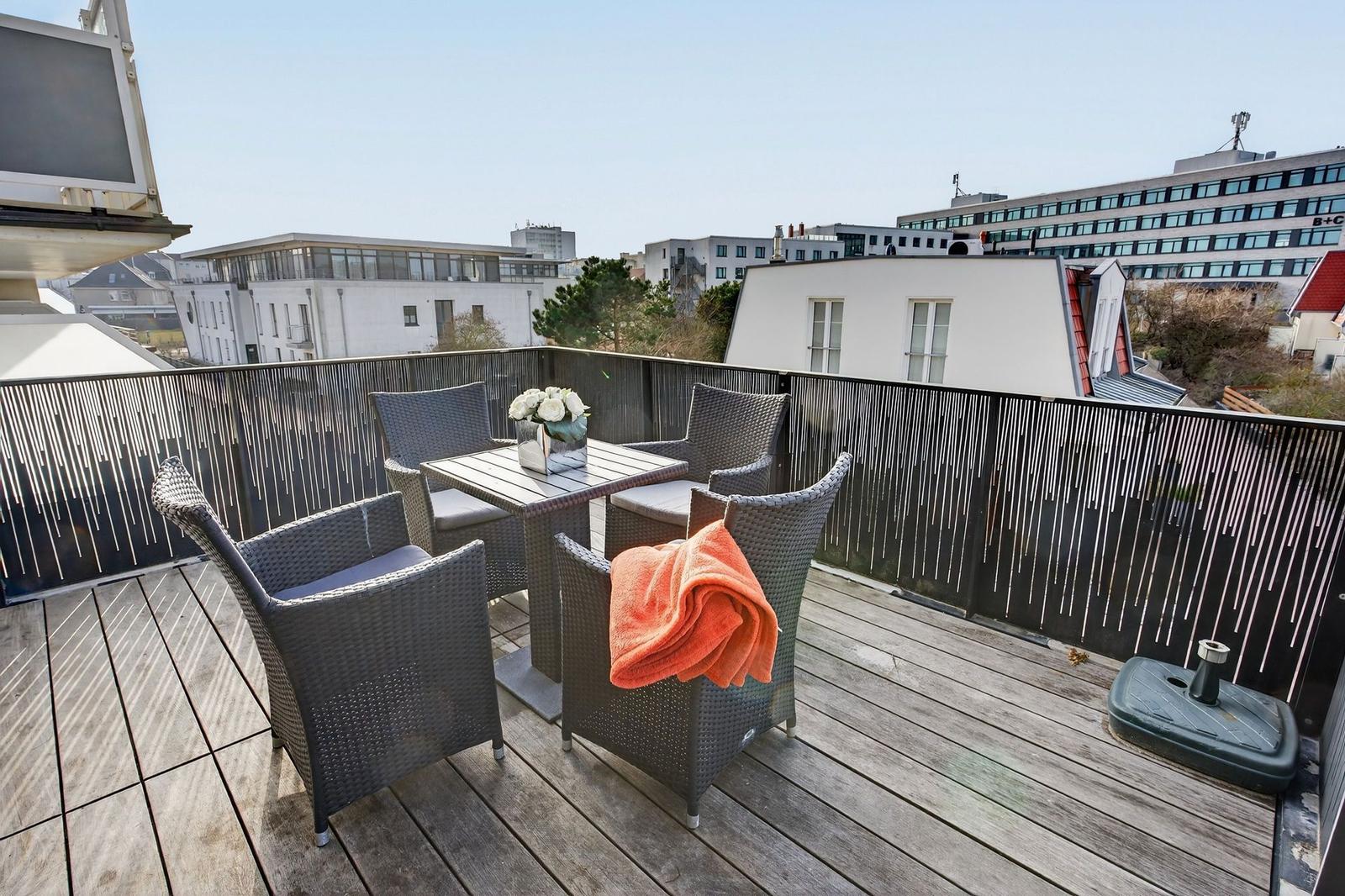 Terrasse mit Tisch, vier Stühlen und orangefarbener Decke. Blick auf städtische Umgebung.