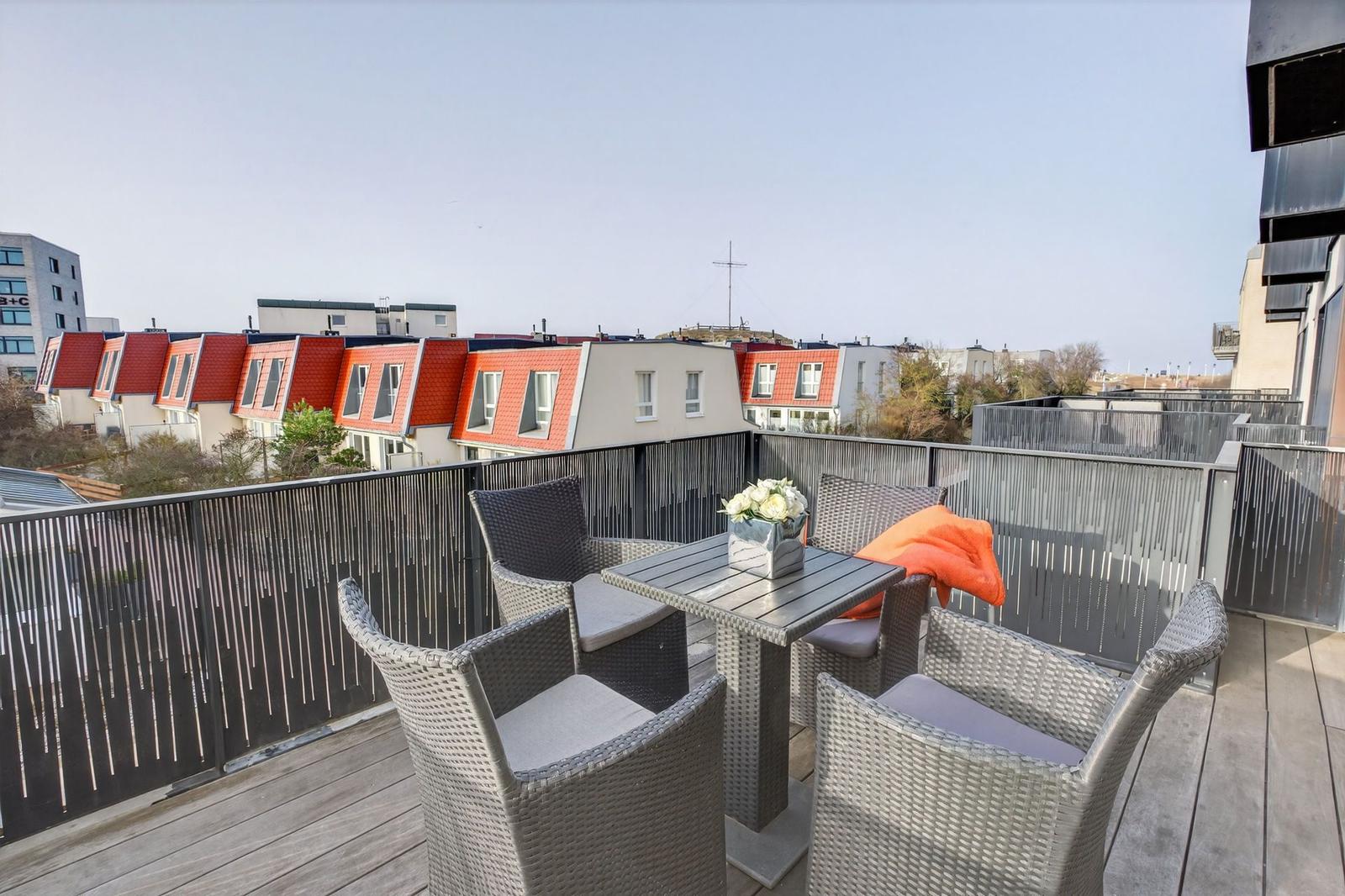 Terrasse mit Tisch und vier Stühlen, Blick auf rote Dächer und Stadt.