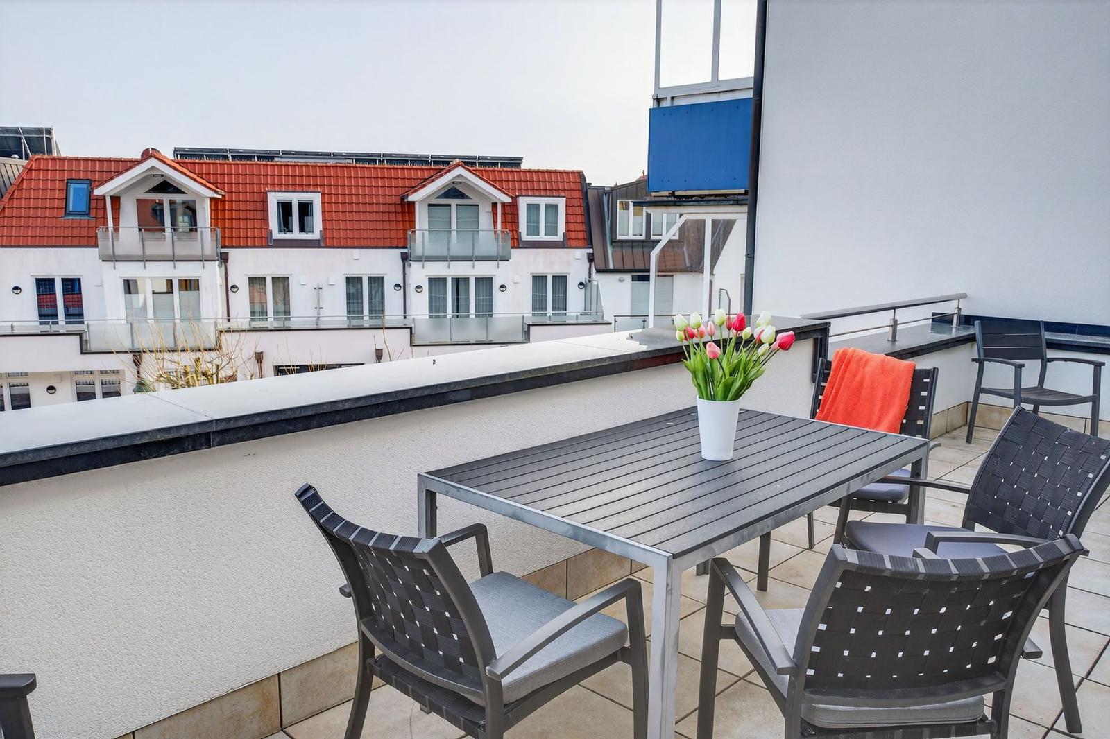 Terrasse mit Tisch, Stühlen und Blumen. Blick auf Wohnhäuser mit roten Dächern.