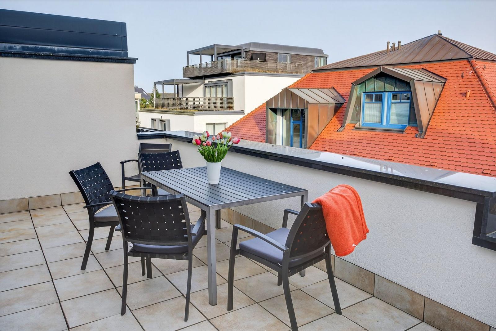 Terrasse mit Tisch, Stühlen und Blumen. Blick auf Dächer und Fenster.