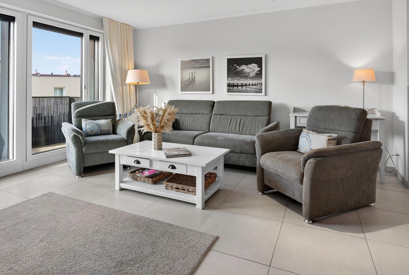 Wohnzimmer mit grauer Couch, zwei Sesseln, weißem Tisch und Balkon.