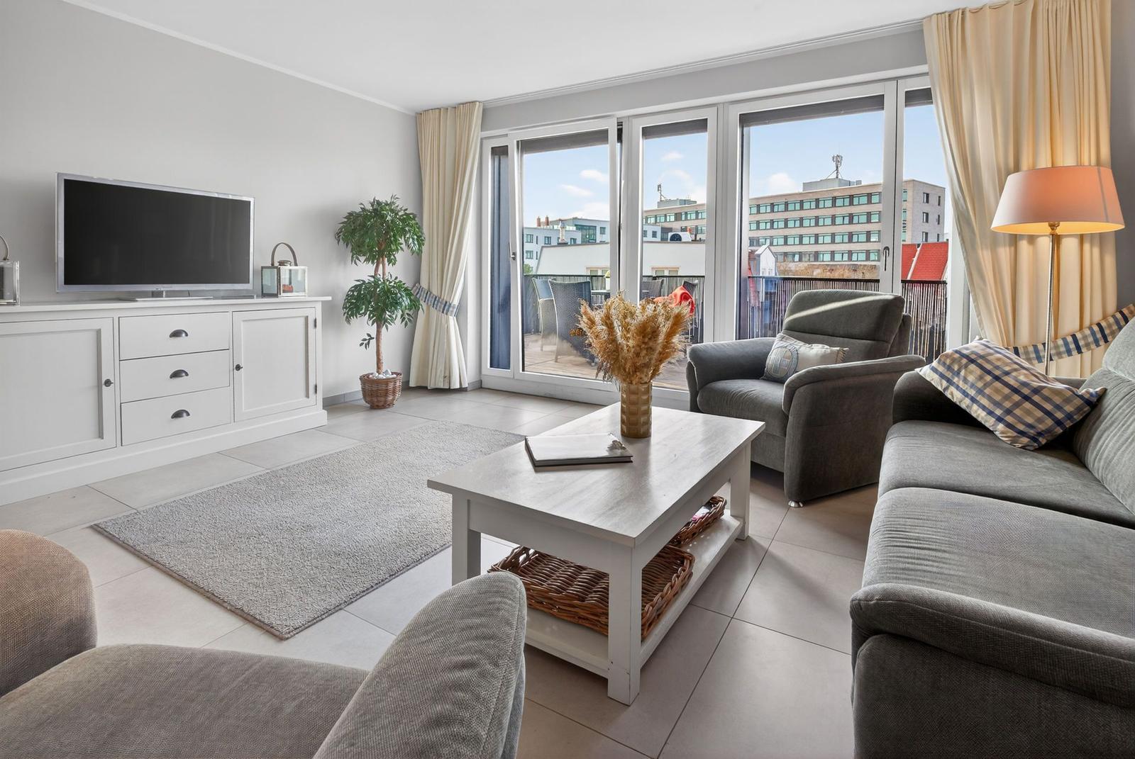 Wohnzimmer mit Sofa, Sessel und Fernseher. Balkon mit Blick auf Gebäude.