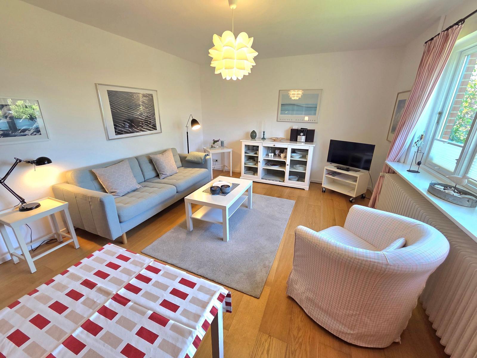 Wohnzimmer mit Sofa, Sessel, Couchtisch und Fernseher. Holzboden und große Fenster.