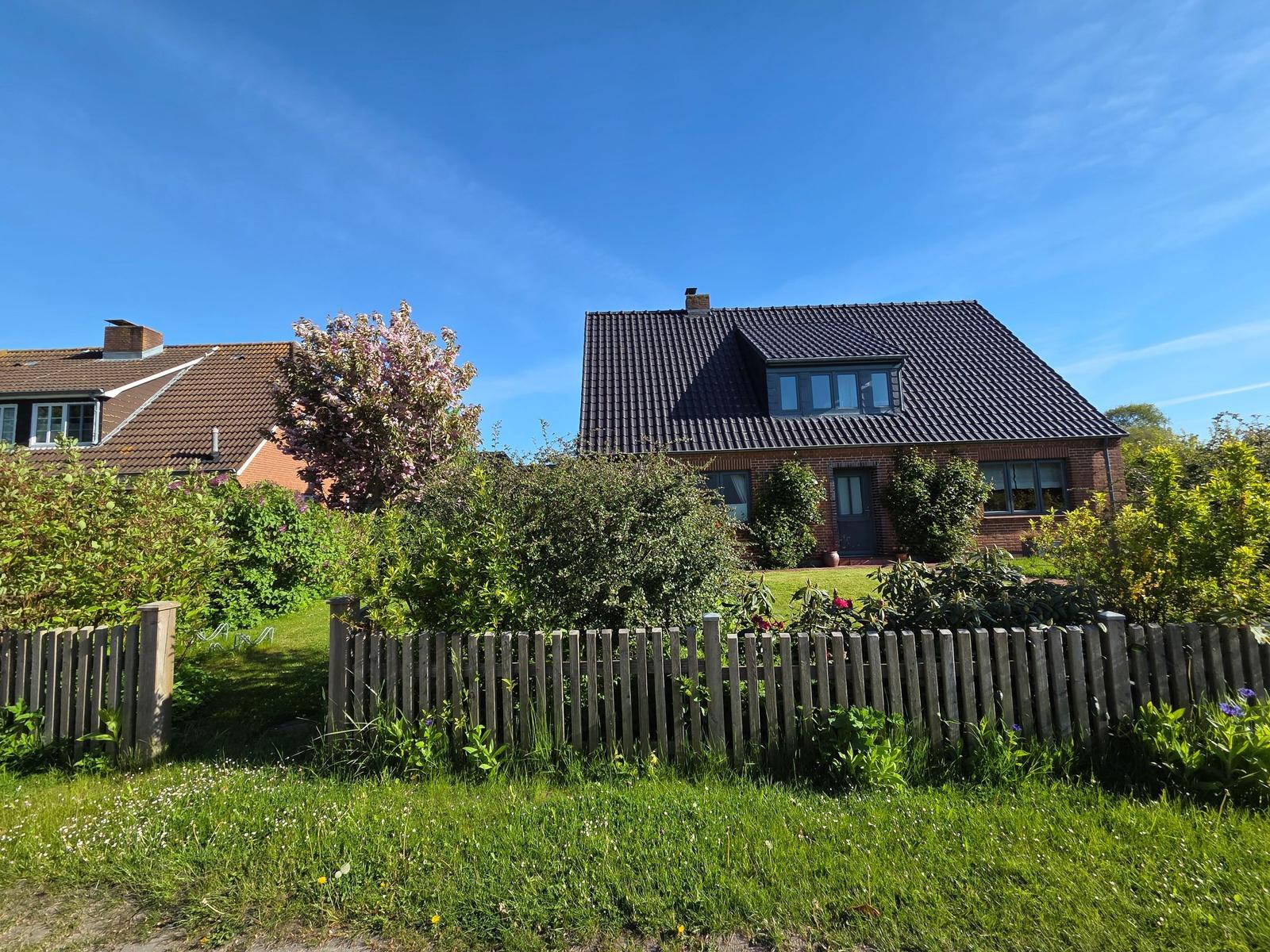 Haus mit Gartenzaun, Blumen und Baum unter blauem Himmel.